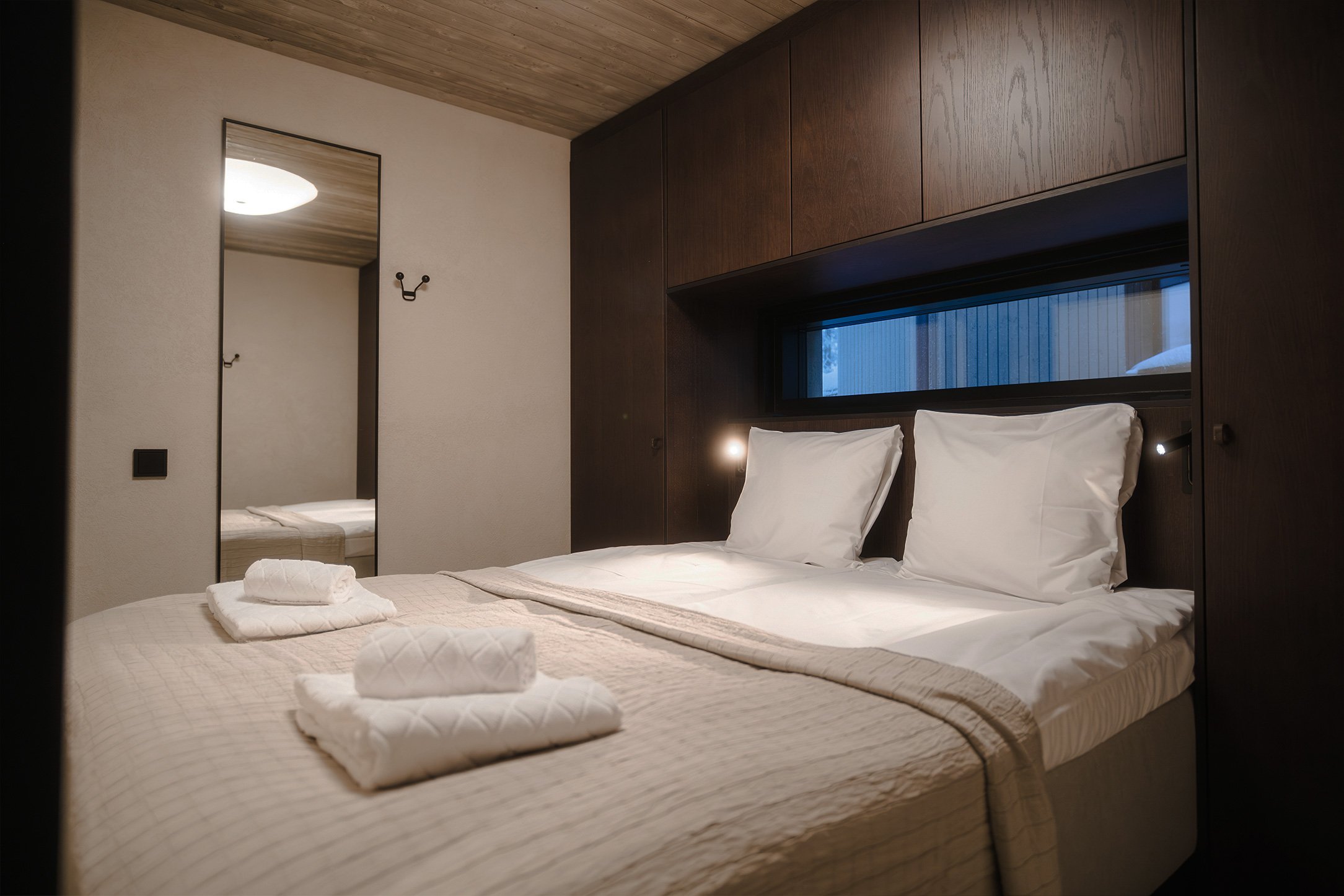 A neatly made bed with white pillows and beige bedding in a modern hotel room. Towels are folded on the bed, and a large mirror and wall hooks are visible in the background.
