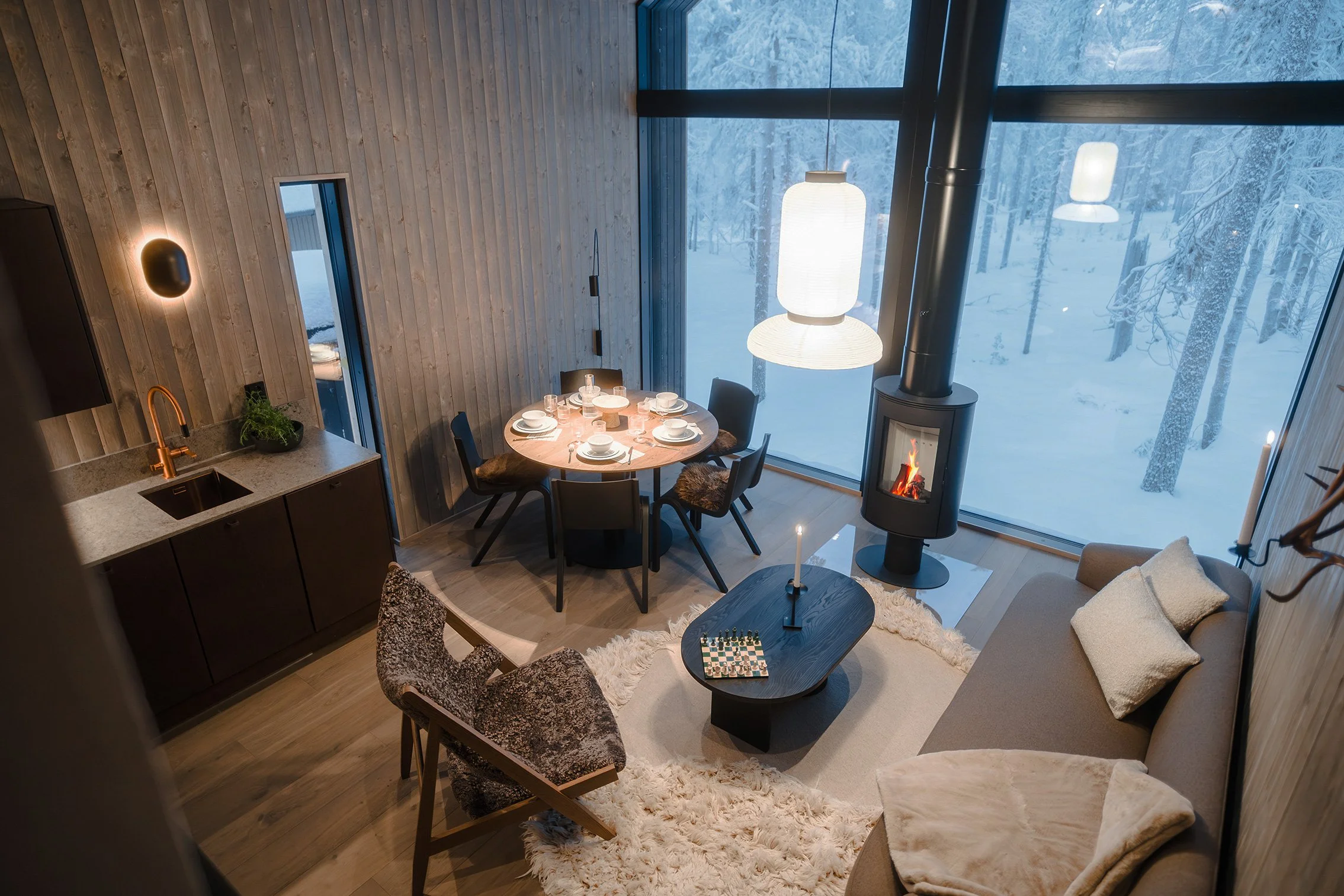 Cozy living and dining area inside a modern cabin with large windows showing snowy landscape, wood-paneled walls, a round dining table set for four, a sofa with throw pillows, a black fireplace, and hanging light fixtures.