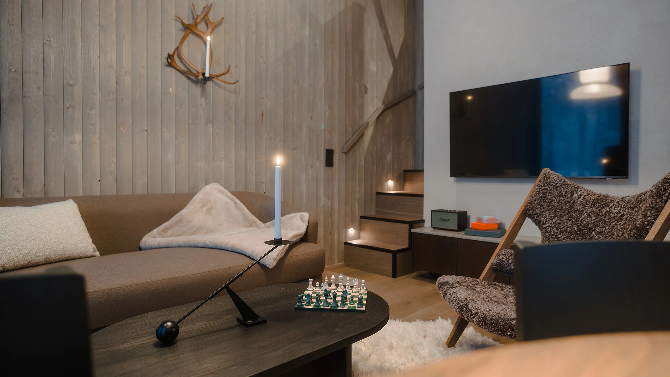 Cozy living room with a beige sofa, a blanket, pillows, a wooden coffee table with a candle, a chessboard, and a telescope. There's a TV on the wall opposite the sofa, wooden staircase with lights, and a wall with wood paneling decorated with antlers