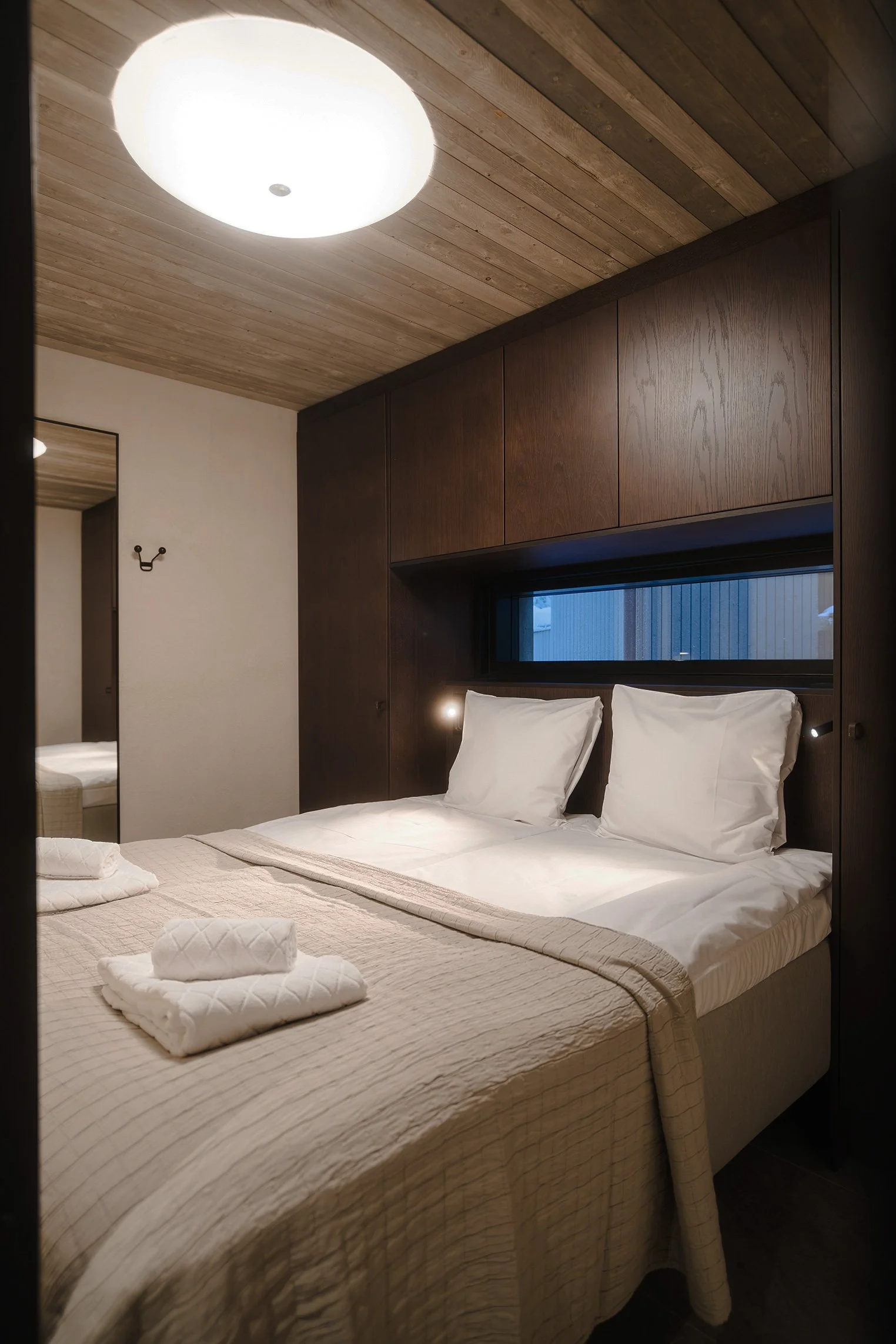 A cozy hotel bedroom with a neatly made bed, white linens, pillows, folded towels, and soft lighting, featuring dark wood accent walls and a wood-paneled ceiling.