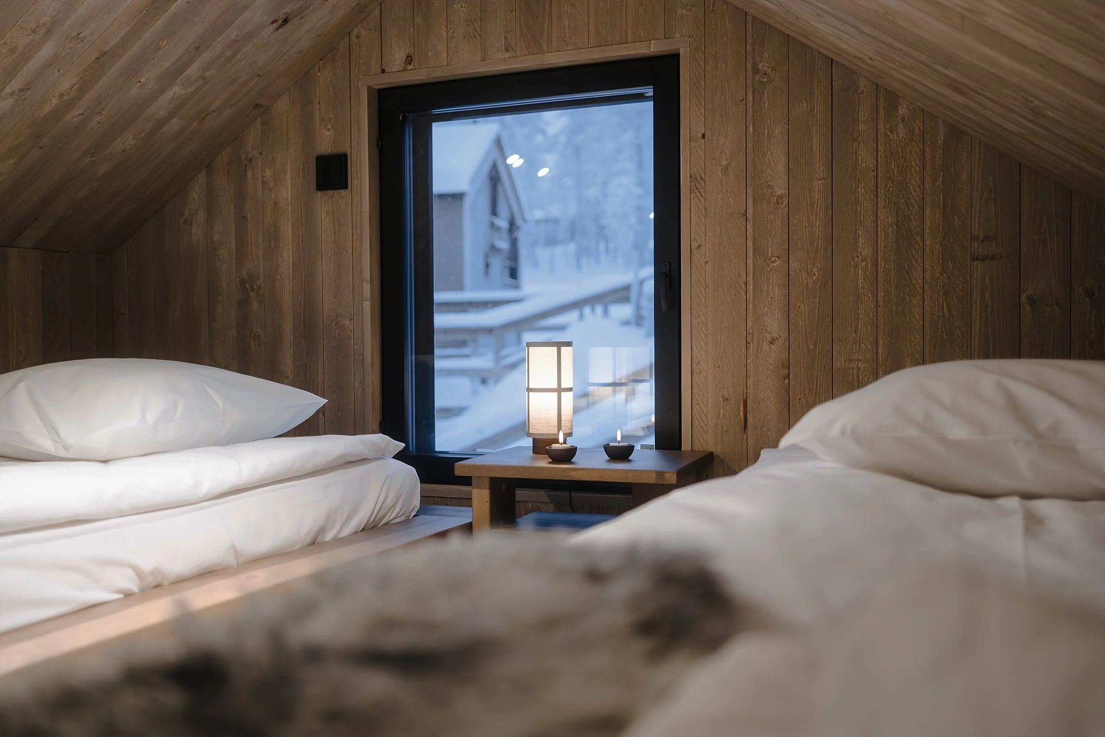 Cozy bedroom with wooden walls, two beds with white bedding, and a window showing a snowy outdoor scene; small table with a lamp and two candles.