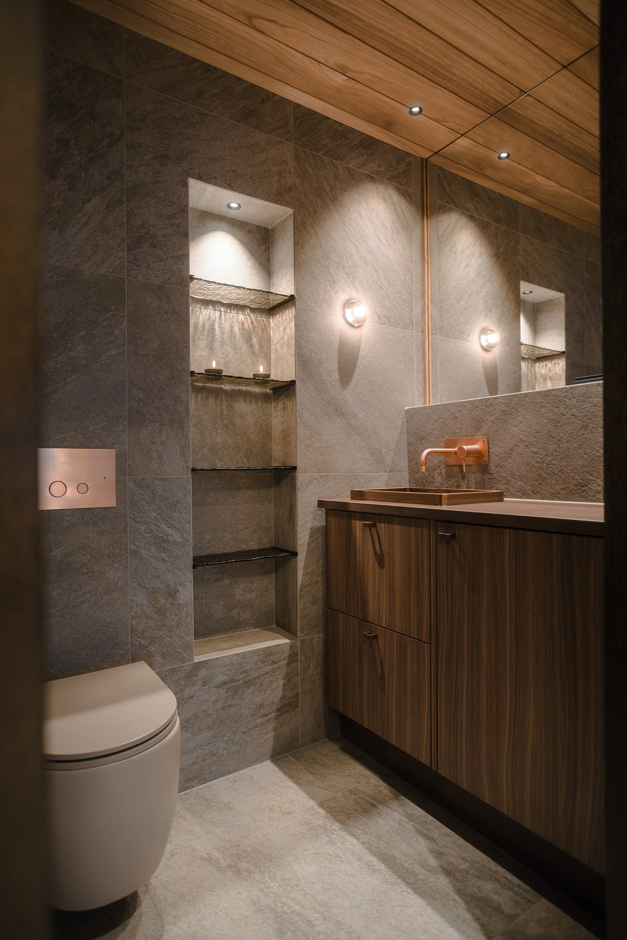 Modern bathroom with grey tiled walls, a wooden ceiling, sleek vanity with a copper sink and faucet, built-in shelves with candles, and a white toilet.