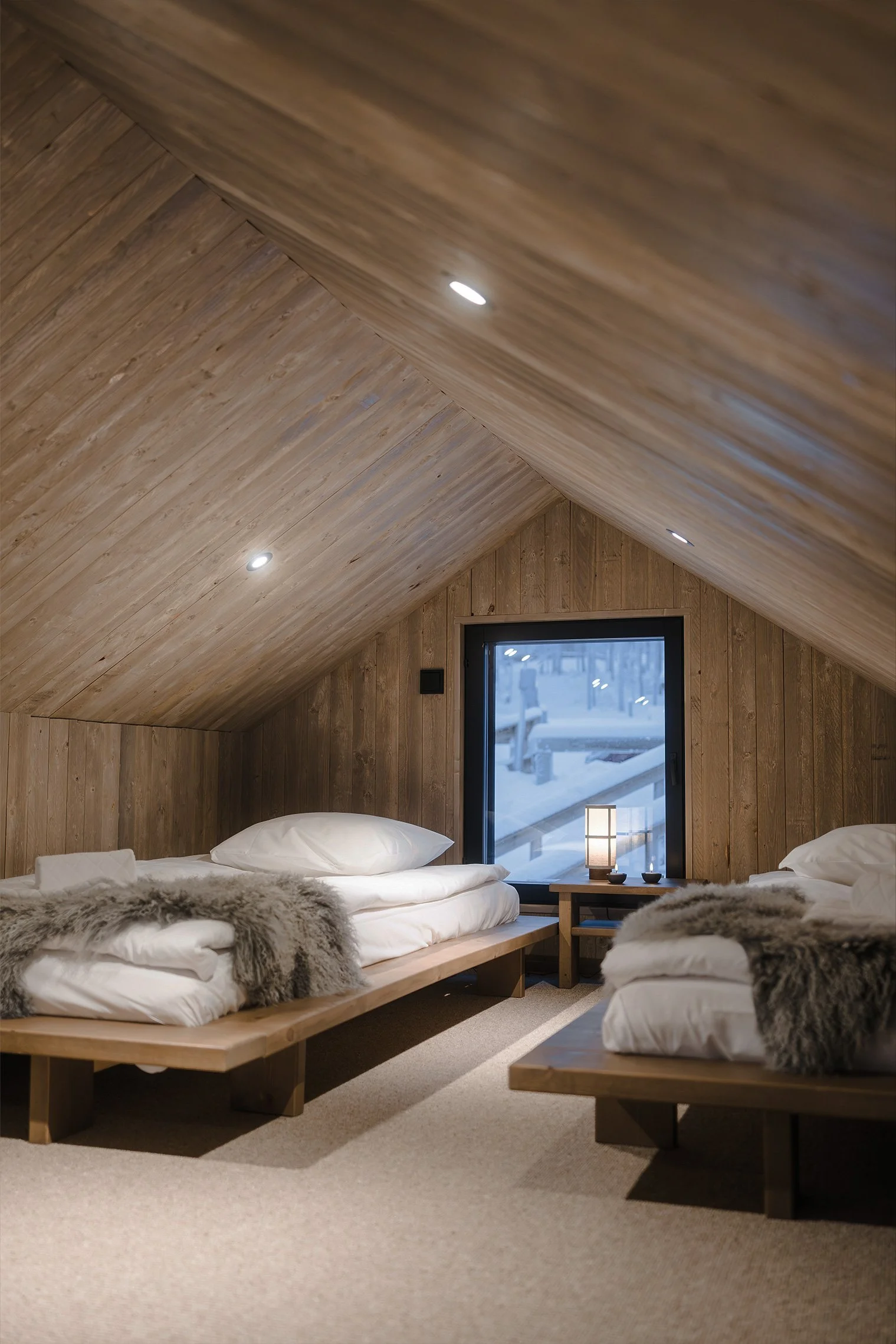 Two beds in an attic bedroom with wood-paneled walls and sloped ceiling, adorned with fur throws, a window showing a snowy scene outside, and a small table with a lamp between the beds.