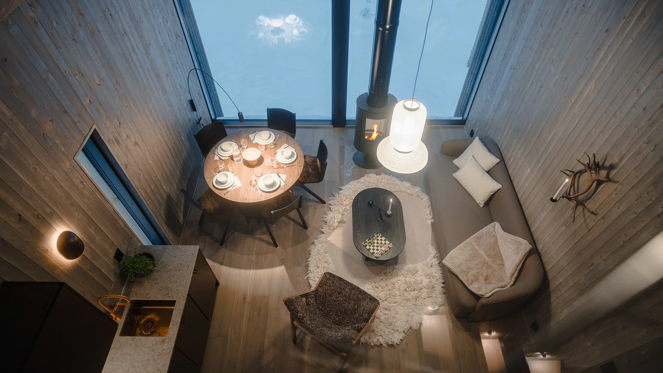 Living room and dining area with a round dining table set for six, a wood-burning stove, a gray sofa with cushions, a shaggy white rug, and a large window showing a snowy outdoor scene.