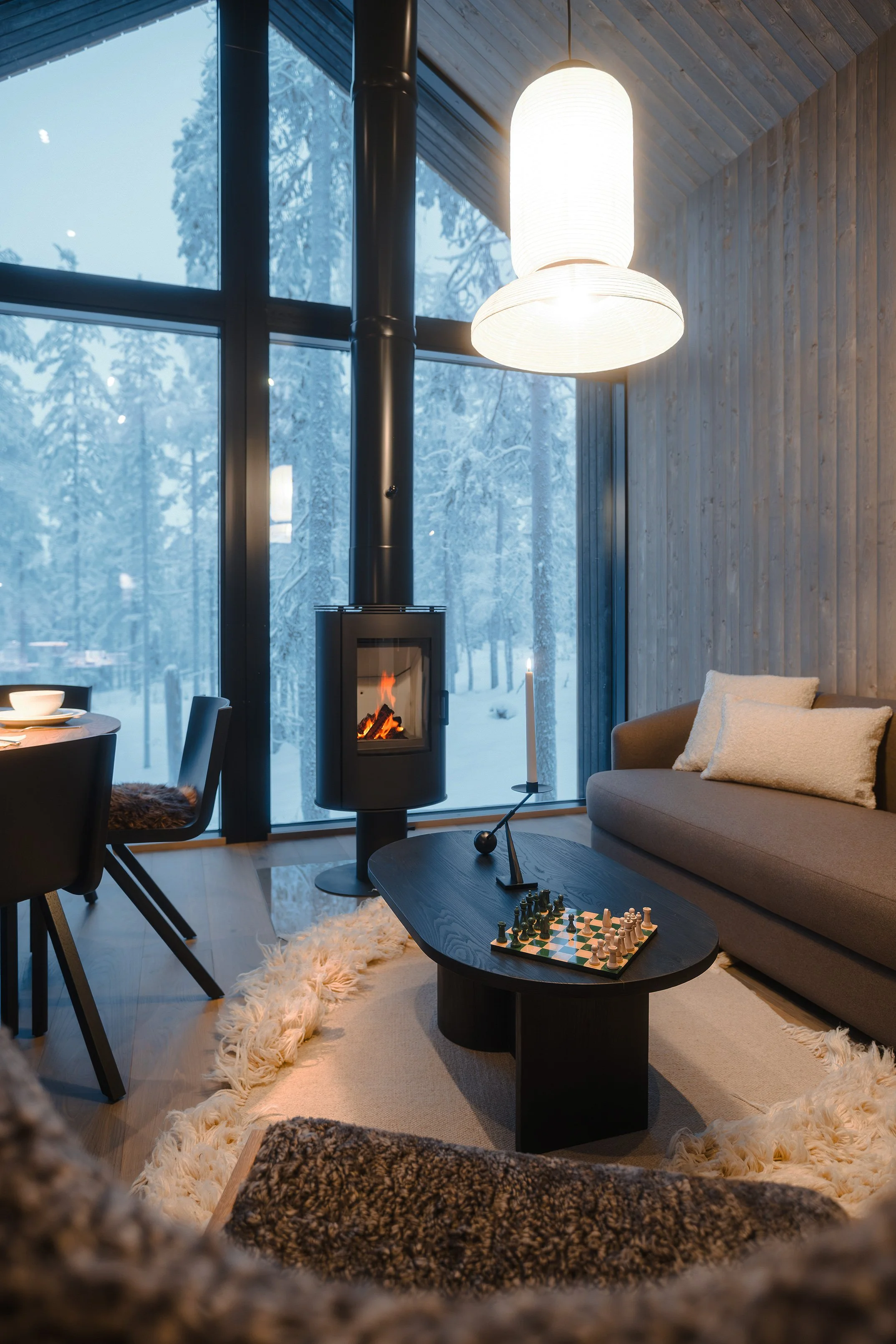 Living room with large snow-covered windows, a wood-burning stove, a gray sofa with pillows, a black oval coffee table with chess set and candle holder, round beige rug, and modern lighting fixtures.