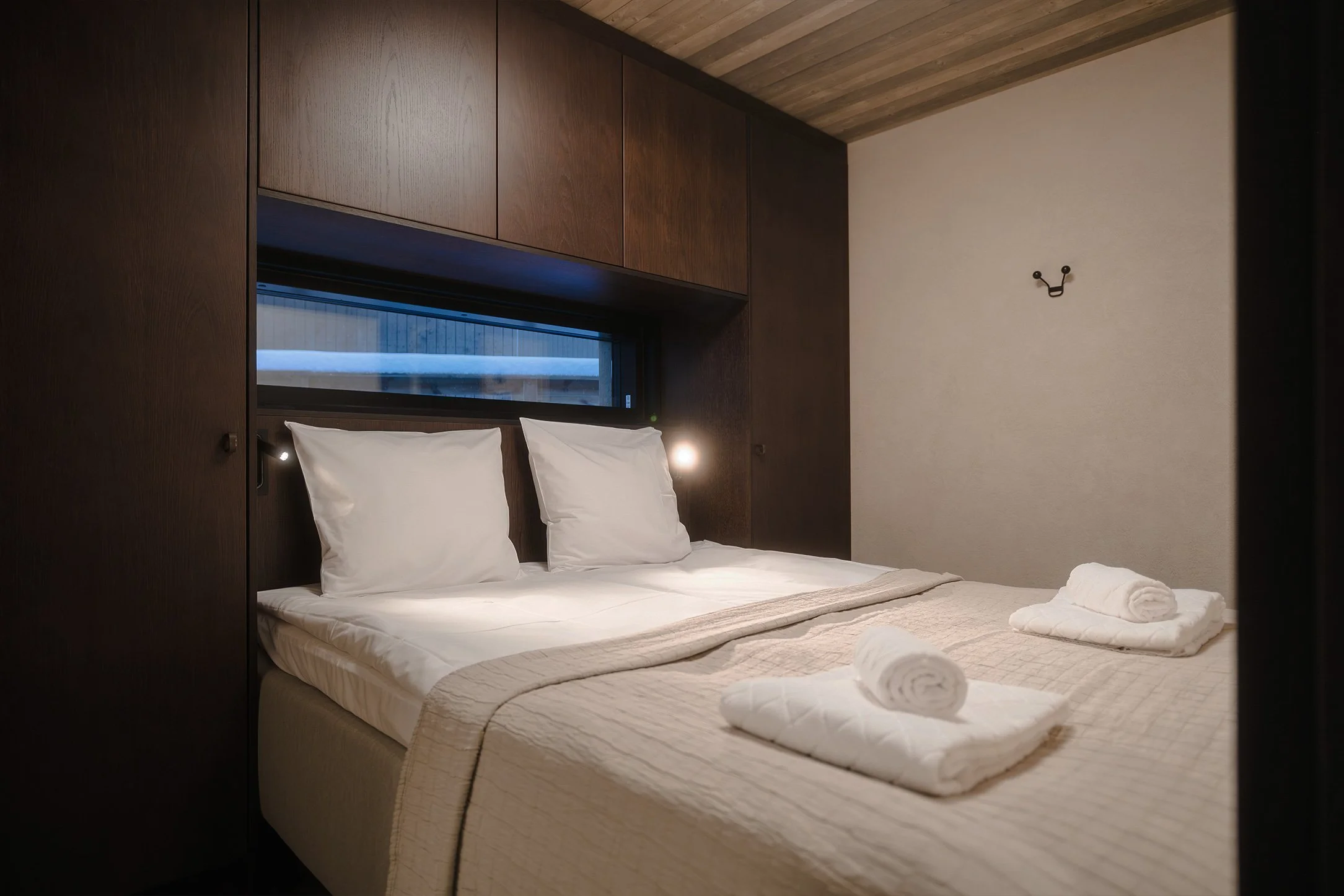 A neatly made bed with white pillows and rolled towels in a modern bedroom with dark wood paneling and a window above the headboard.