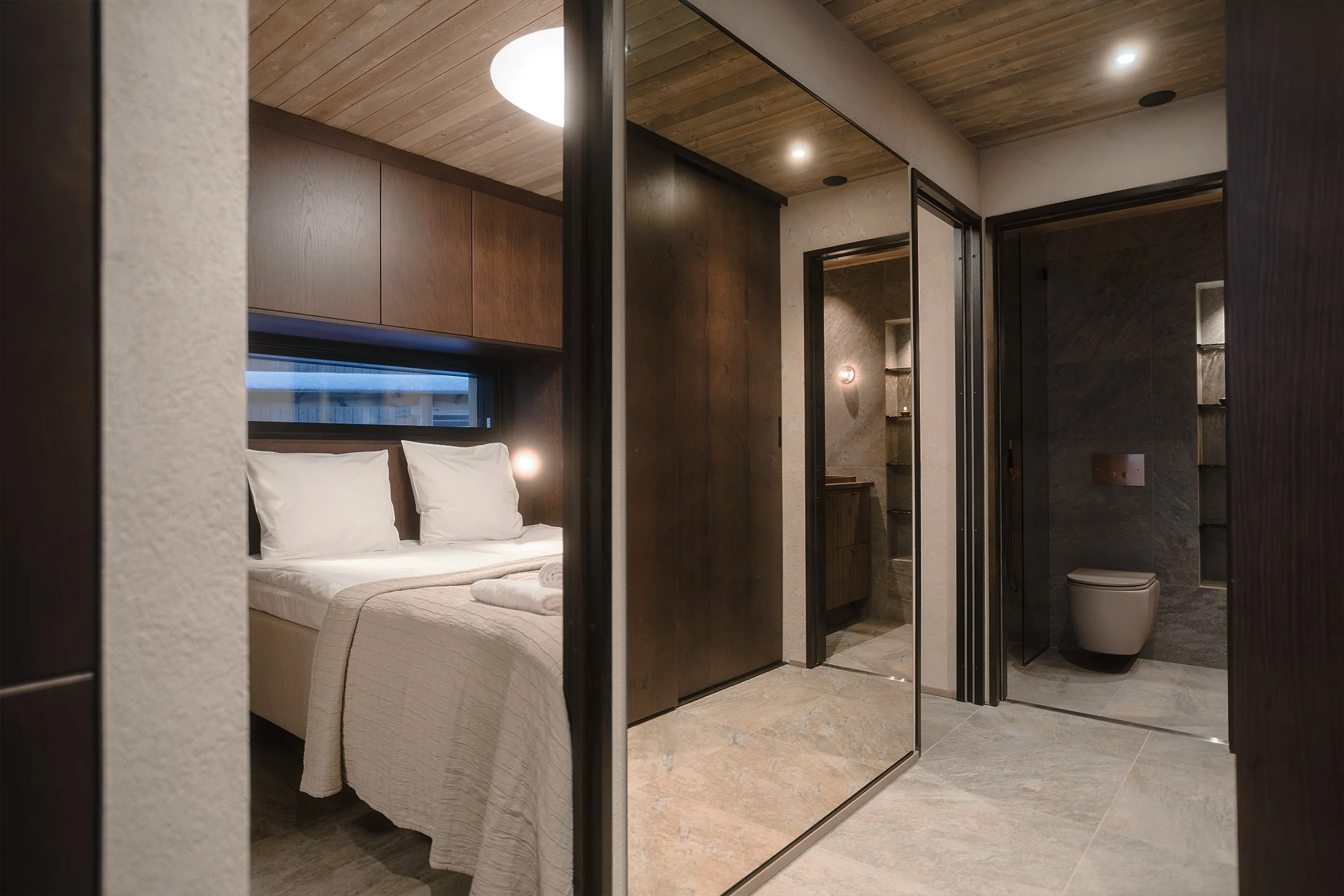 View of a modern hotel room with a bed, a closet with mirror doors, and a bathroom with tiled walls and a toilet.