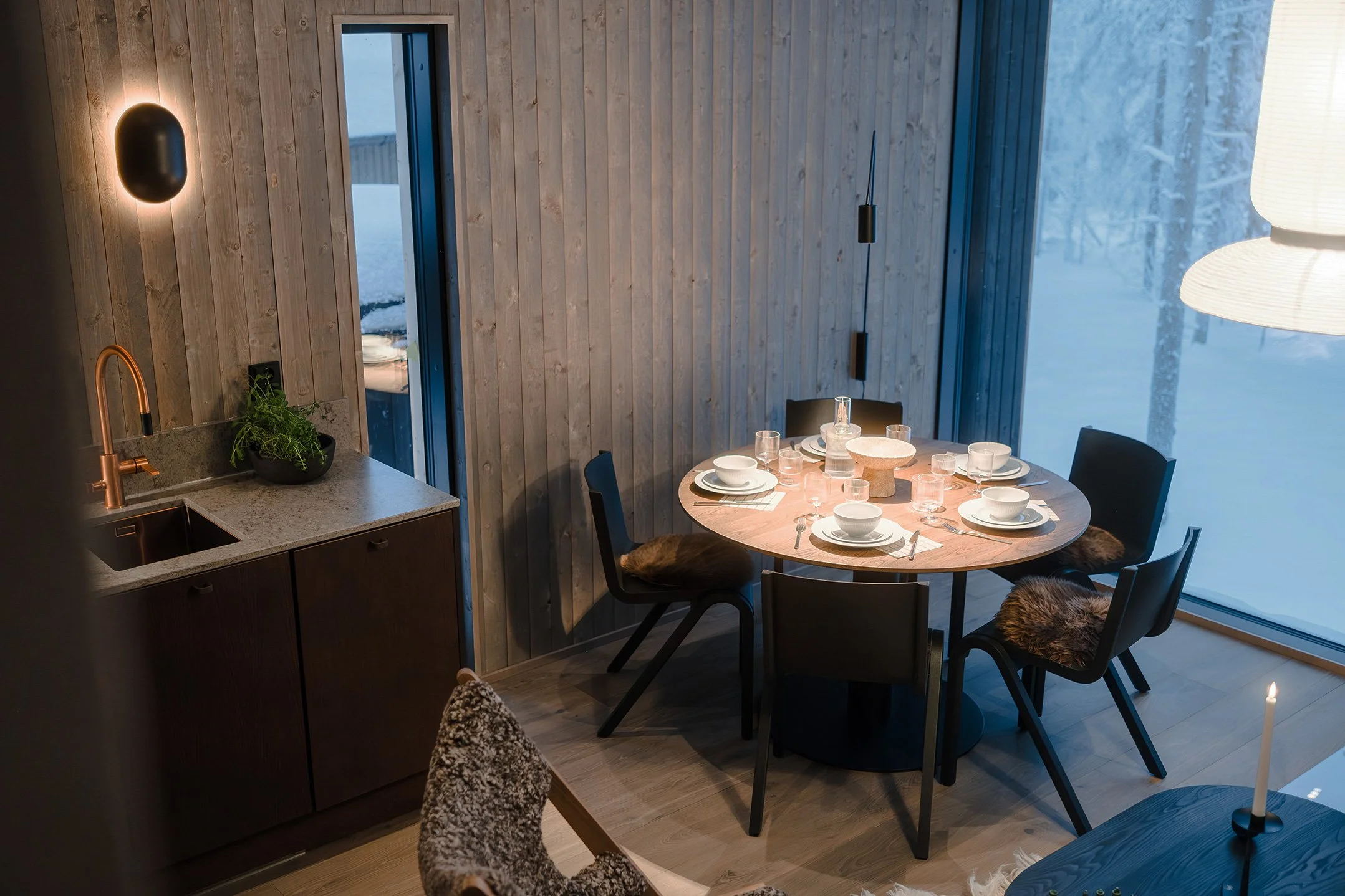 Cozy dining area with a round wooden table set for a meal, surrounded by black chairs with fur seat covers, next to large windows showing a snowy landscape, with a modern wooden wall and a small kitchenette with a plant on the counter.