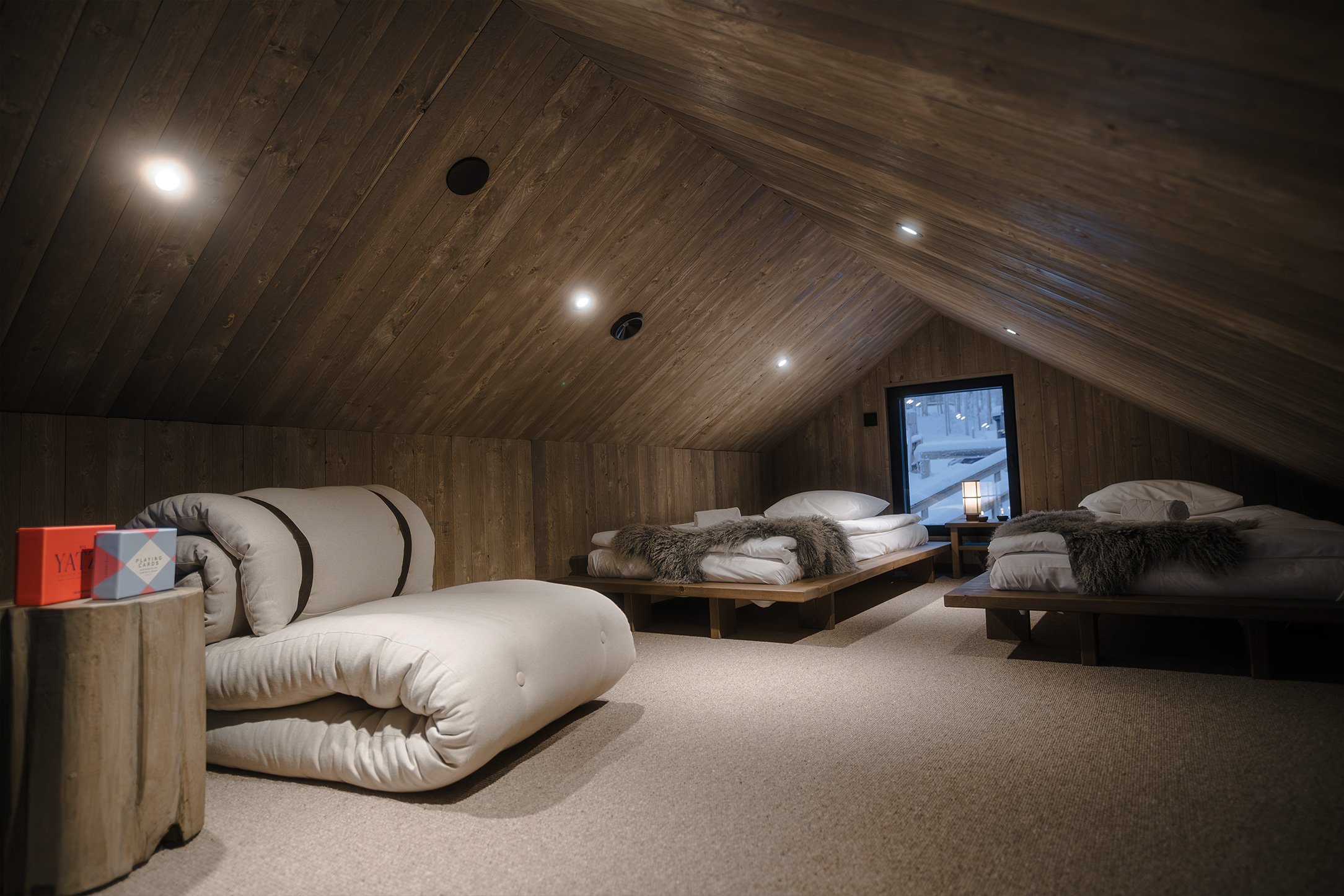 A cozy attic bedroom with three beds, wooden paneling on the sloped ceiling and walls, a small window with a view of snow, a lamp on a nightstand, and plush blankets on the beds.