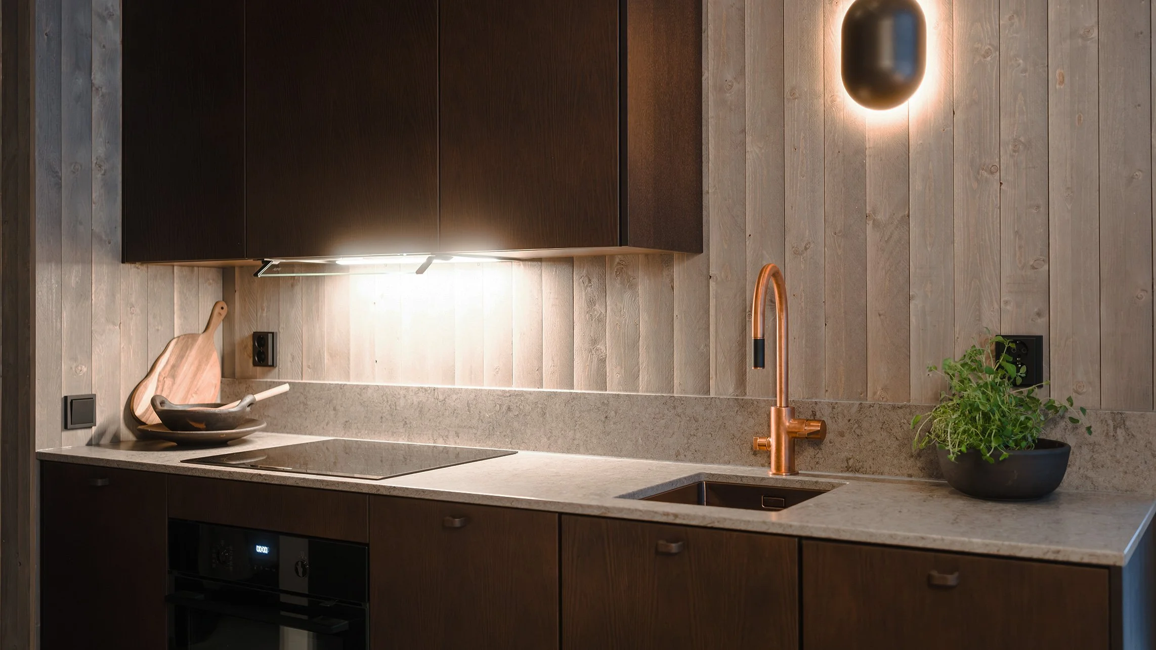 Modern kitchen with dark upper cabinets, light wooden plank wall, a copper faucet, black sink, potted green plant, and stone countertop.