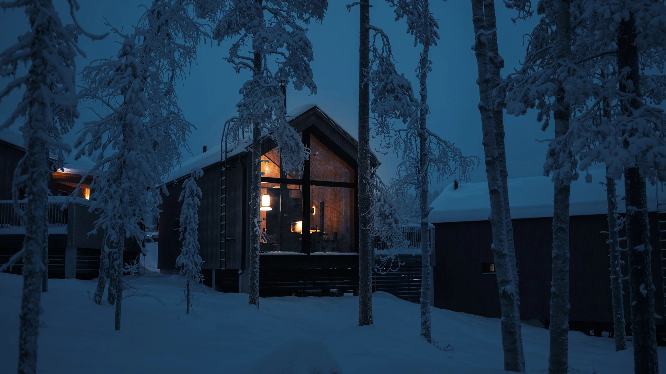 A cozy house with large windows showing warm interior lighting, surrounded by snow-covered trees at dusk.