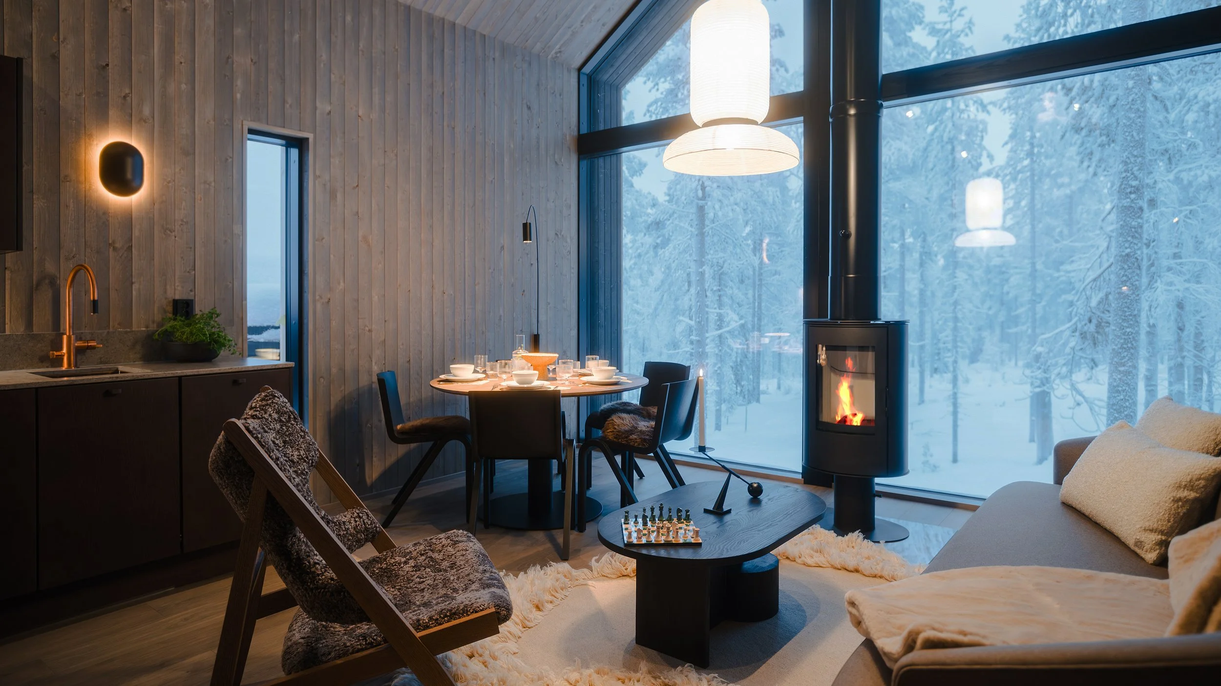 Cozy living room with wood-paneled walls, large window with snowy landscape outside, modern fireplace, dining table set for a meal, and comfortable seating with a chess game on the coffee table.