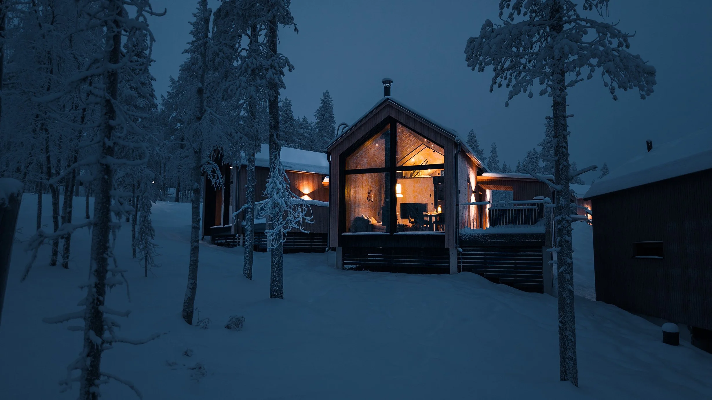 A cozy house illuminated from within, surrounded by snow-covered trees during nighttime with a dark blue sky.