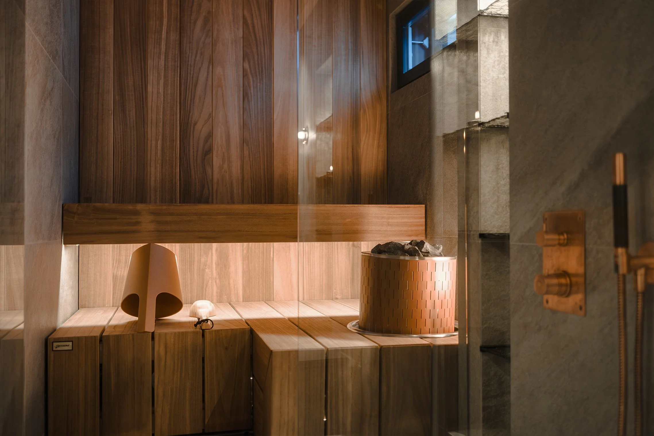Sauna room with wooden benches, a chimney, and rocks for steaming, with a window and modern fixtures.