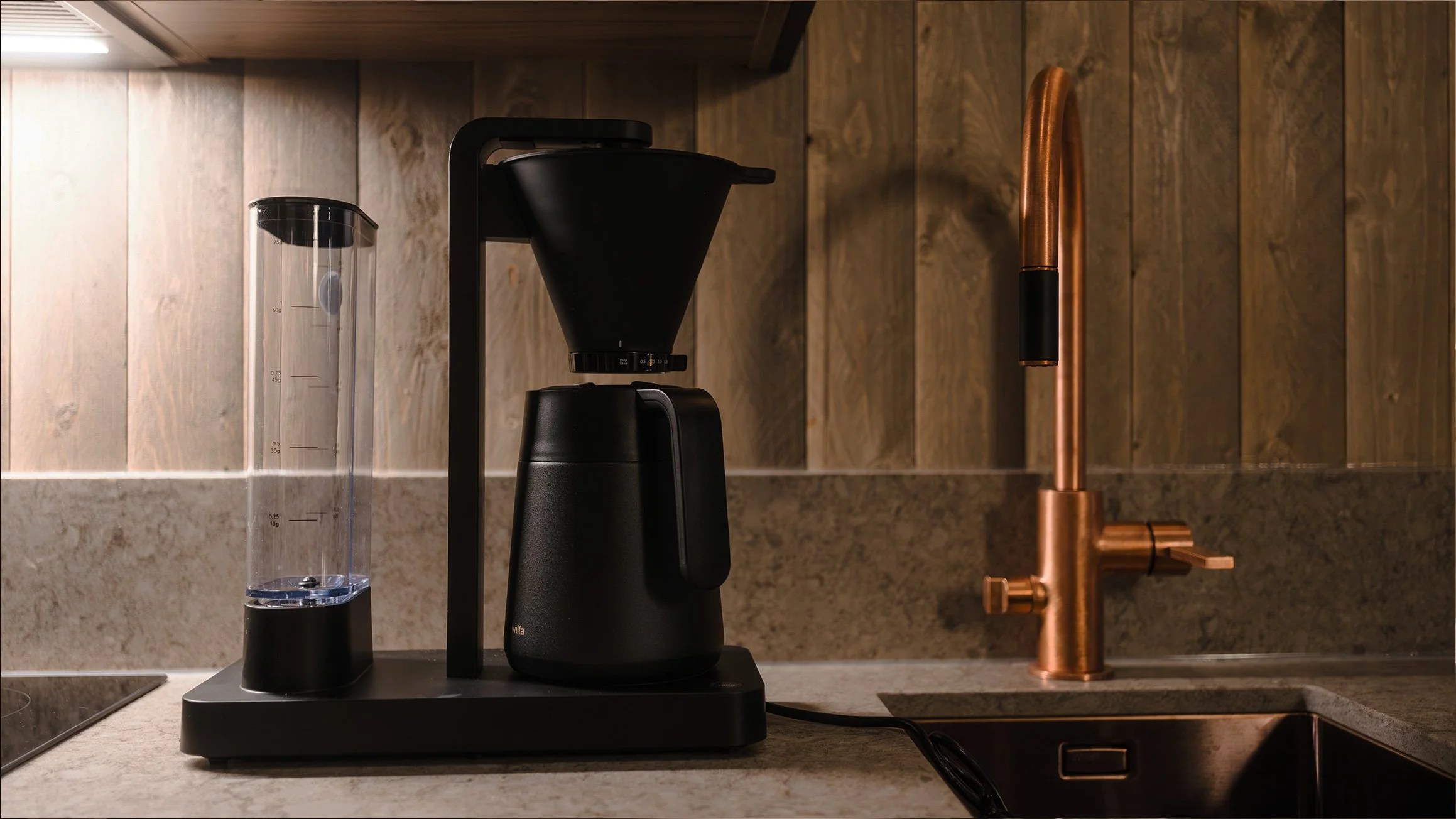 Coffee maker and water pitcher on kitchen countertop with copper sink and wooden backsplash.