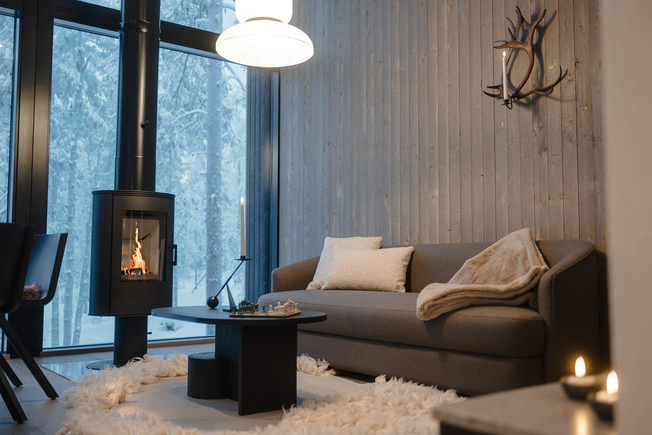 A cozy living room with a gray sofa, decorative pillows, and a blanket, next to a wood-burning stove with a fire, large window showing snow-covered trees outside, a white overhead lamp, and a wall decorated with antler-shaped candle holders.