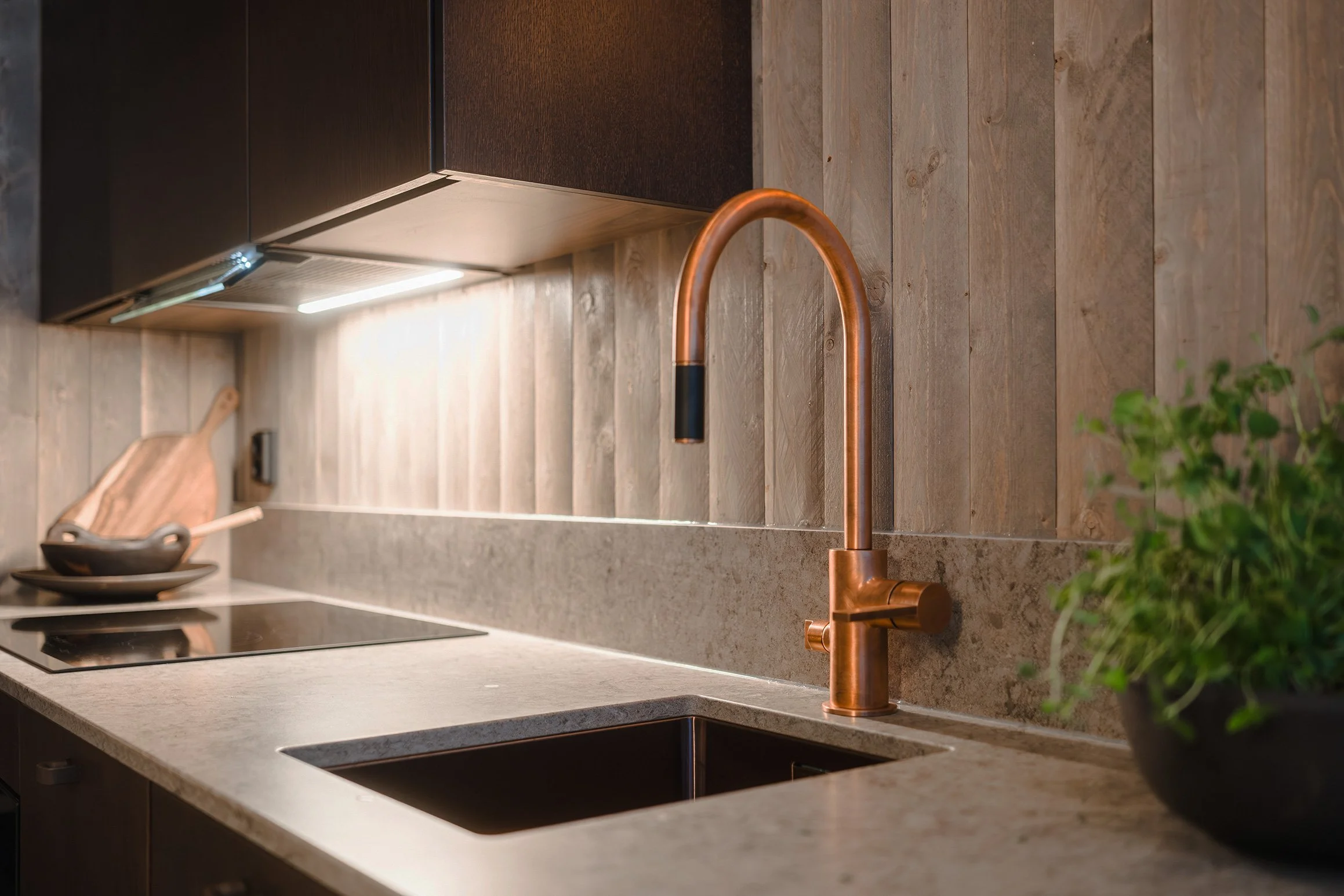 A modern kitchen countertop with a copper faucet over a black sink, a potted plant, a cutting board, and a stovetop with a pot and bowl, with wooden wall paneling and cabinetry.