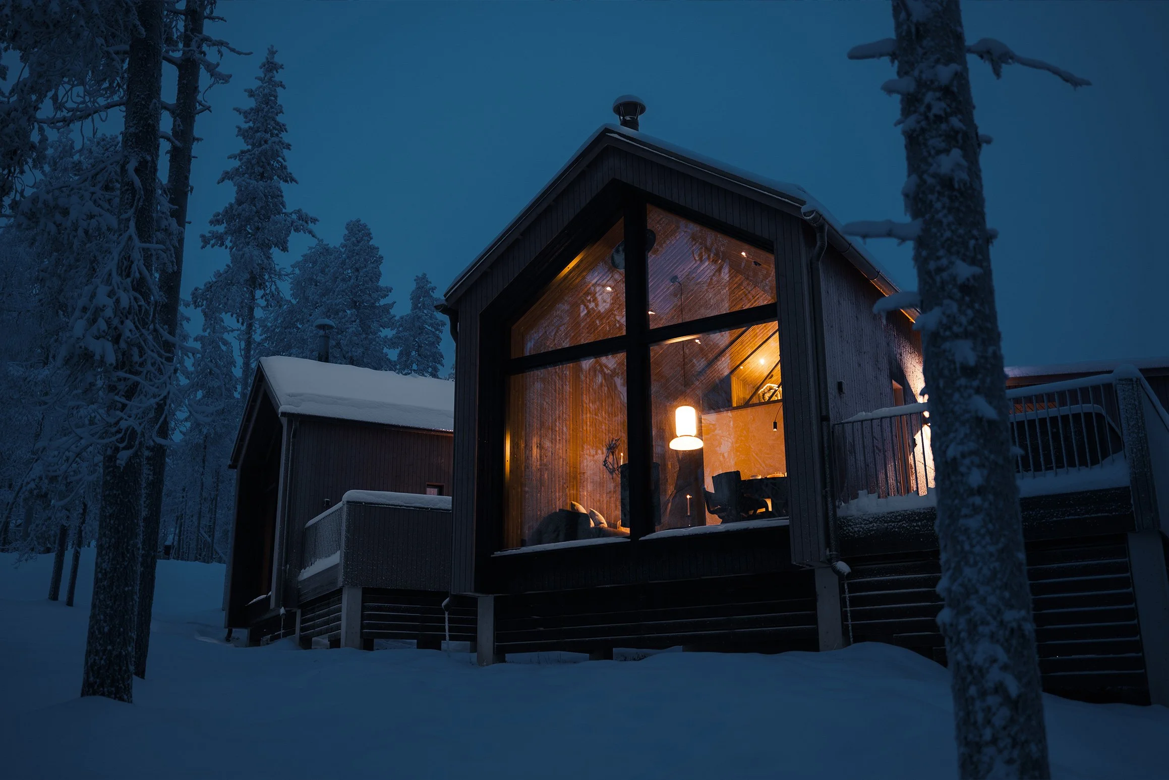 A cozy wooden house with large glass windows glowing with warm light, surrounded by snow and tall snowy pine trees at dusk or night.