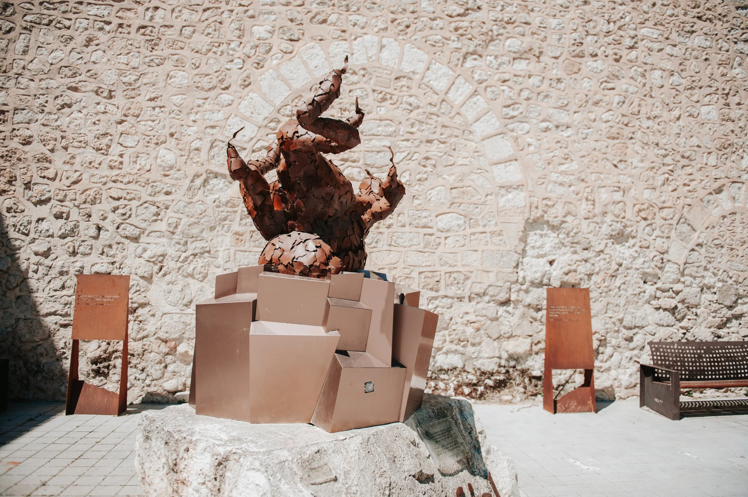 Escultura de un ciervo en un parque, en un pedestal de bloques de metal, con una pared de piedra en el fondo y bancos de madera y señalizaciones alrededor.