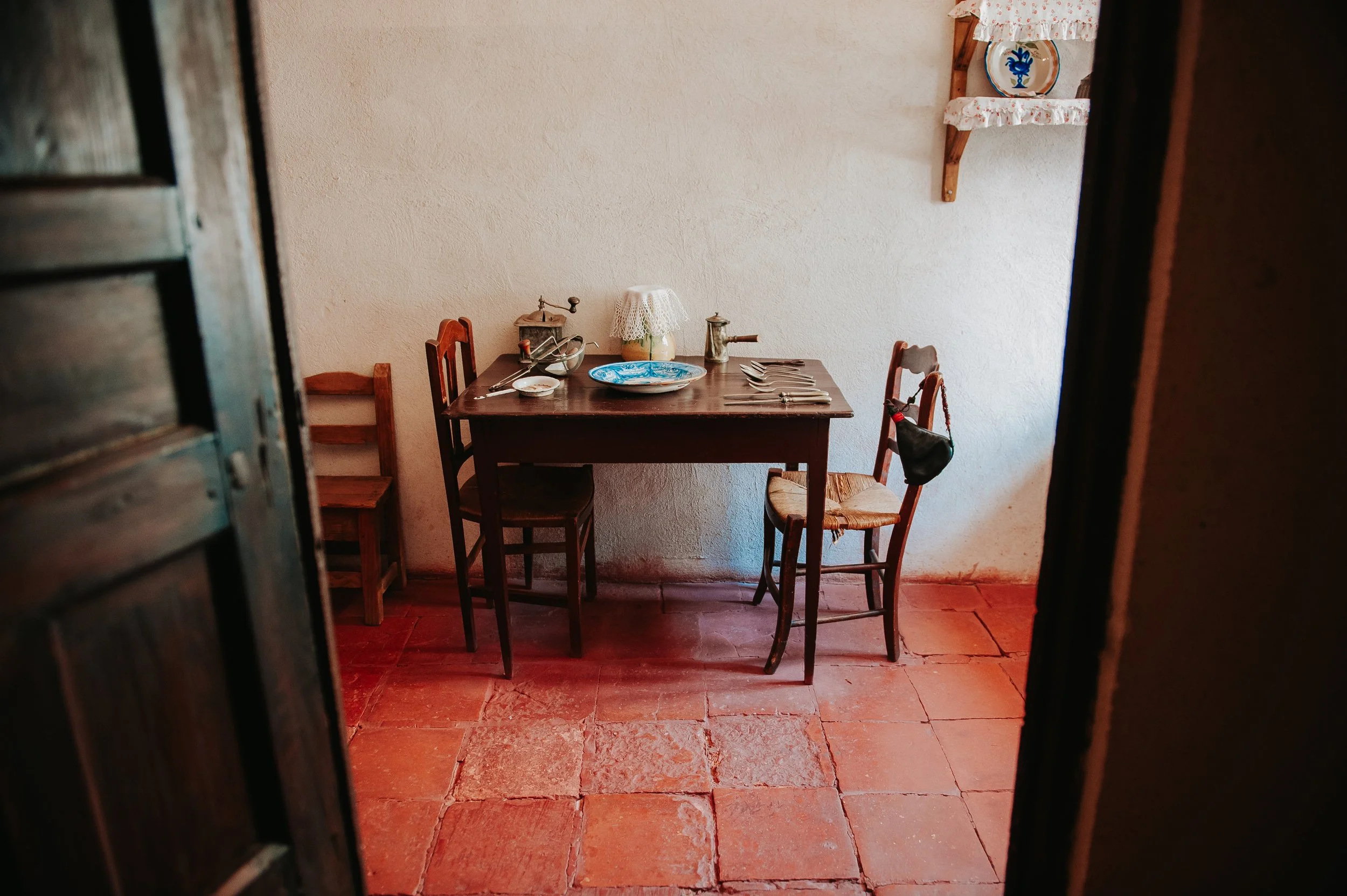 Una mesa de madera con cuatro sillas en una cocina antigua, decorada con utensilios y vajilla tradicional, en un ambiente rústico con paredes de color claro y suelo de terracota.