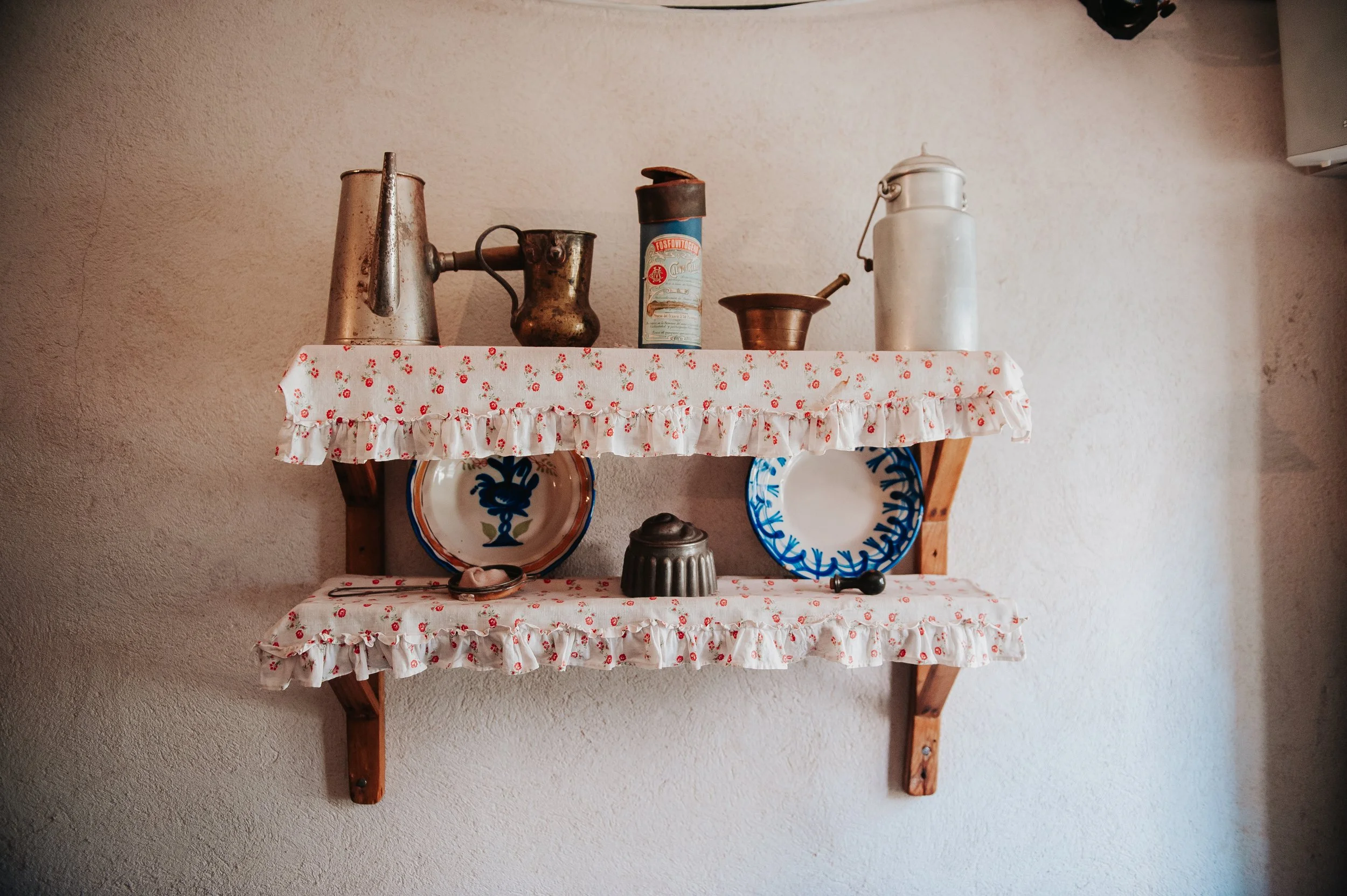 Estantes de madera con adornos de tela con rosas, que sostienen platos de cerámica decorativa, teteras y otros utensilios de metal antiguos.