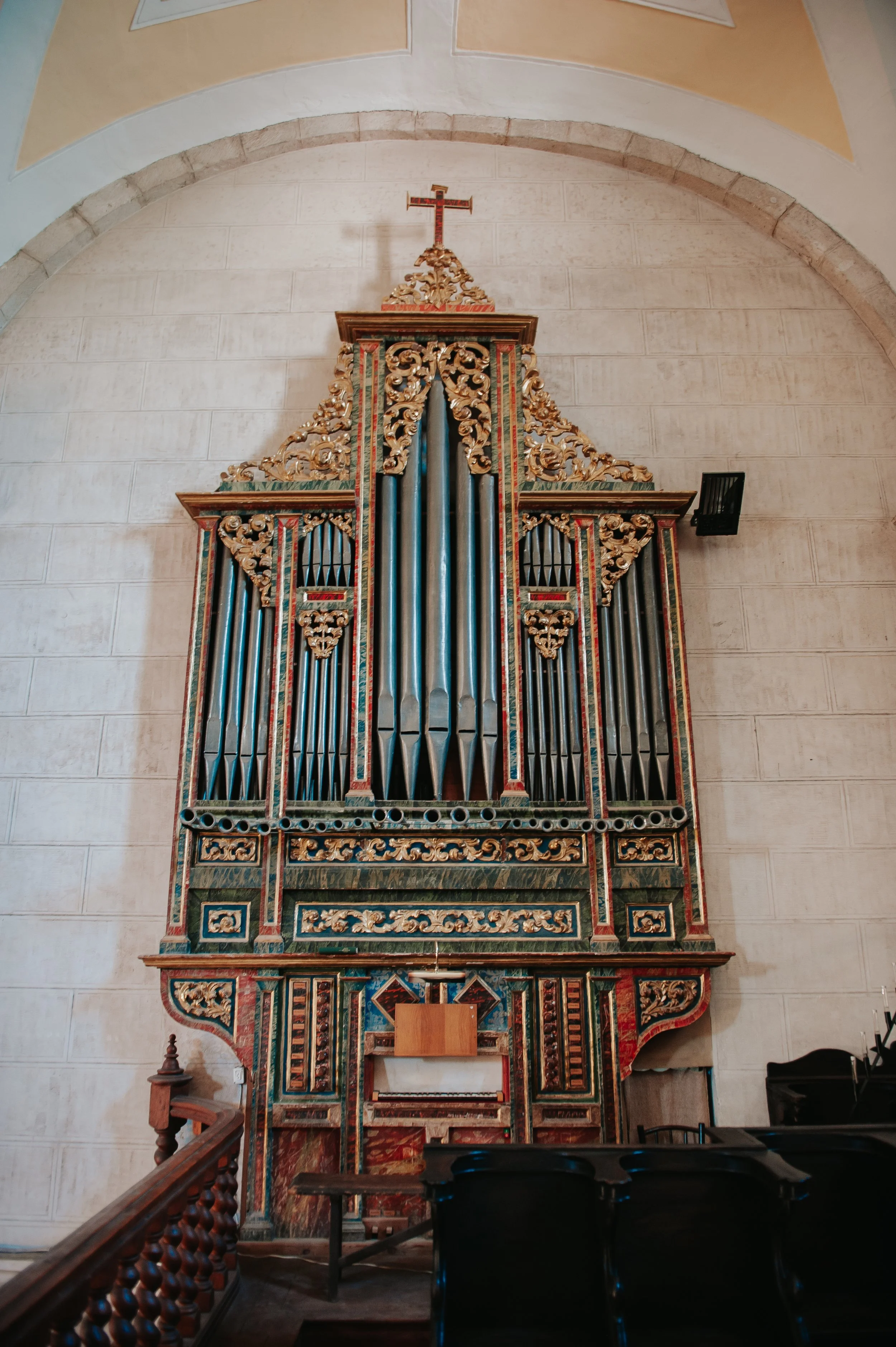 Órgano de iglesia decorado con detalles dorados y madera pintada en colores rojo y verde, con una cruz en la parte superior, ubicado en el interior de una iglesia.