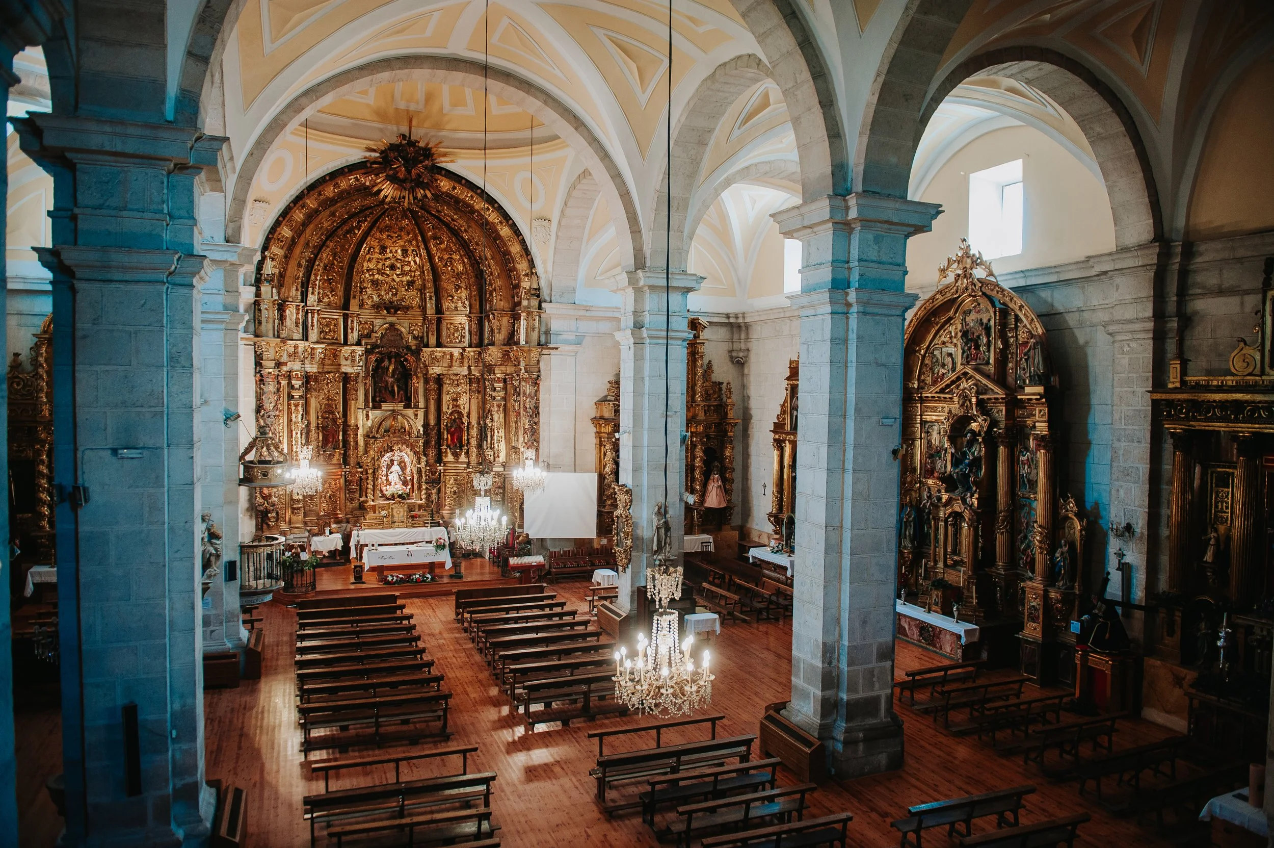 Interior de una iglesia decorada con altares dorados y candelabros, con bancos de madera y columnas de piedra