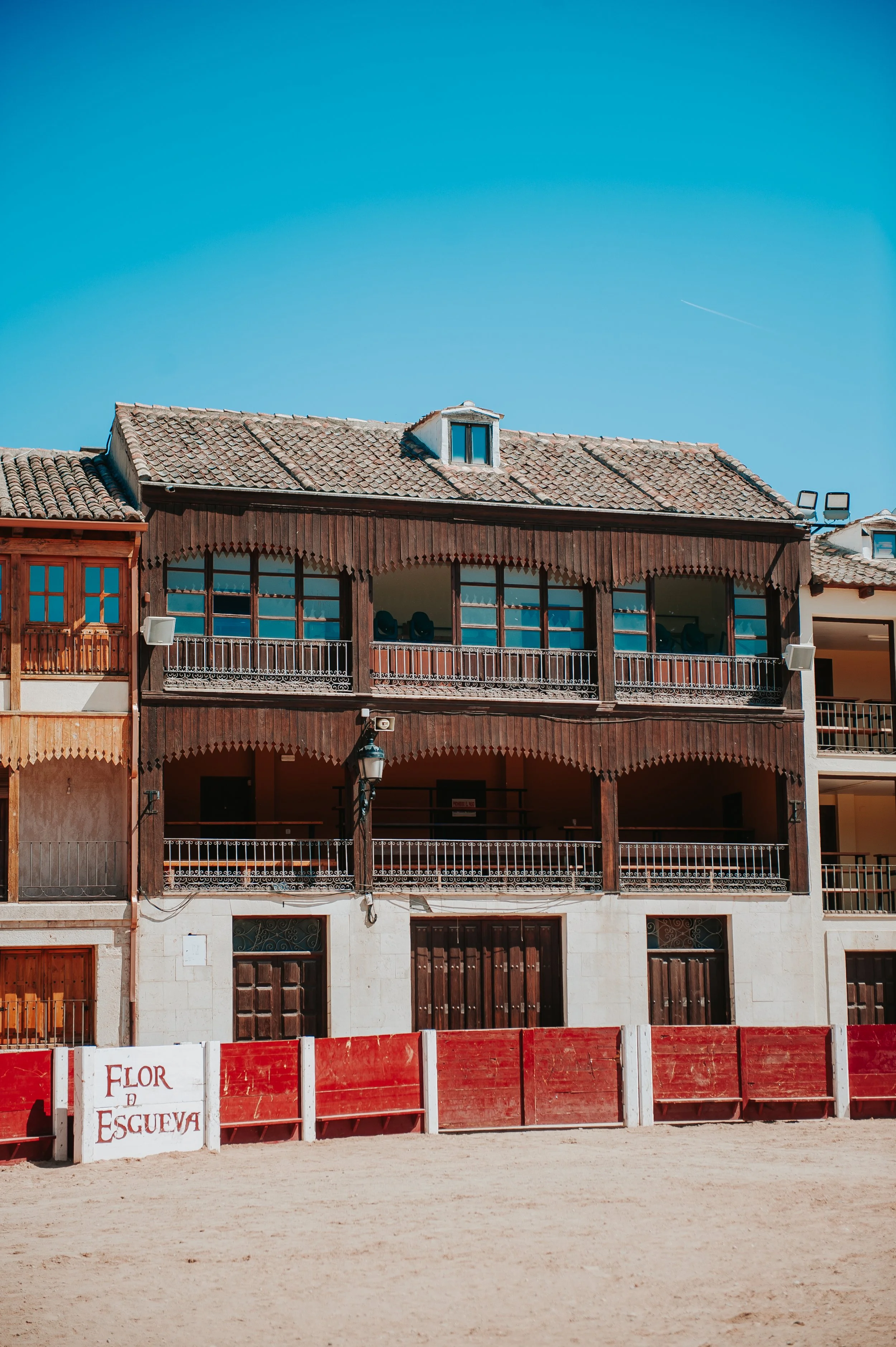 Edificio de madera con balcones de estilo tradicional, en una calle con señal de advertencia y pared roja en primer plano, bajo un cielo azul claro.