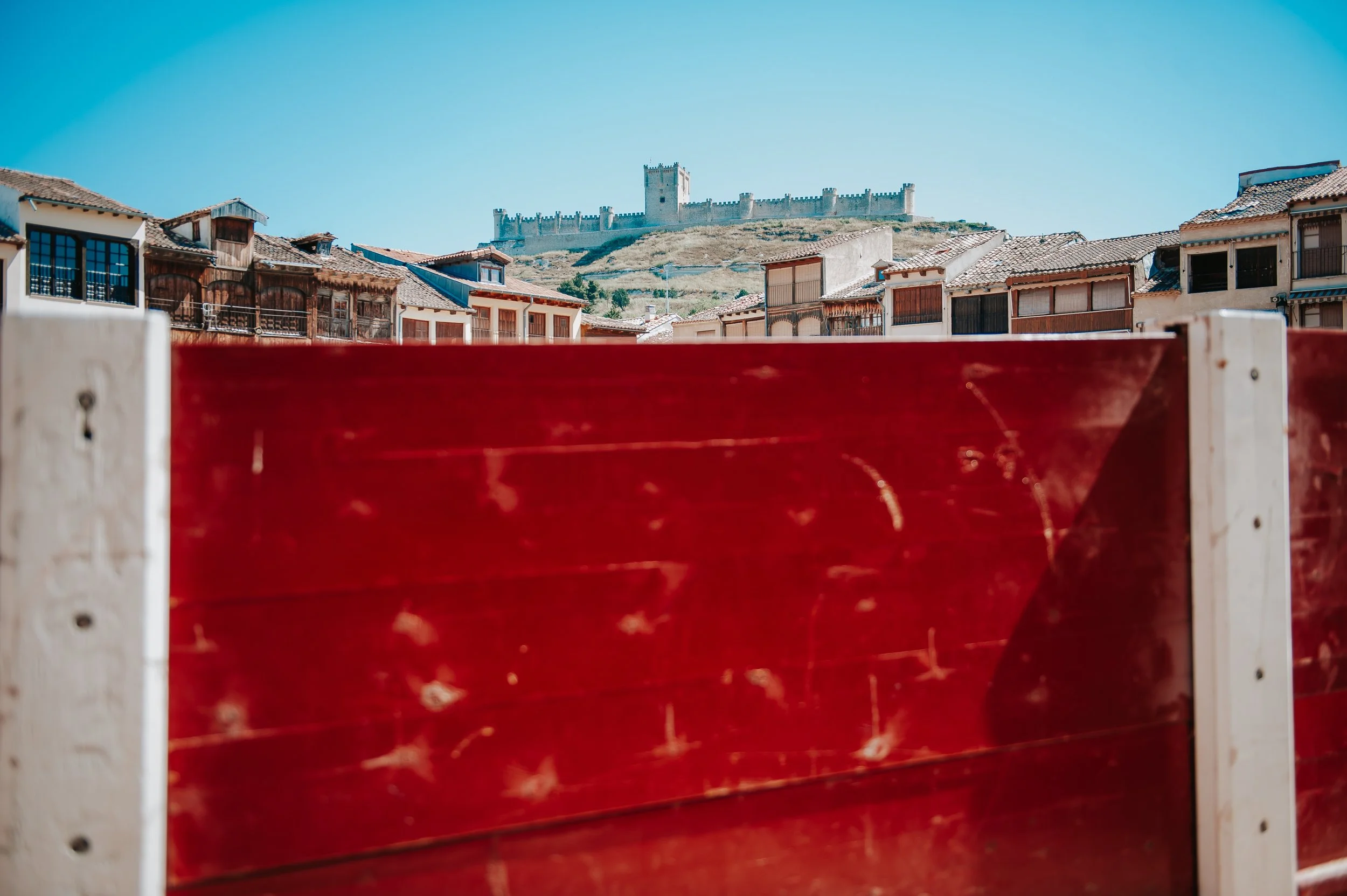 Vista de una ciudad con casas tradicionales y un castillo en una colina al fondo, vista parcialmente bloqueada por una estructura de madera roja en primer plano.