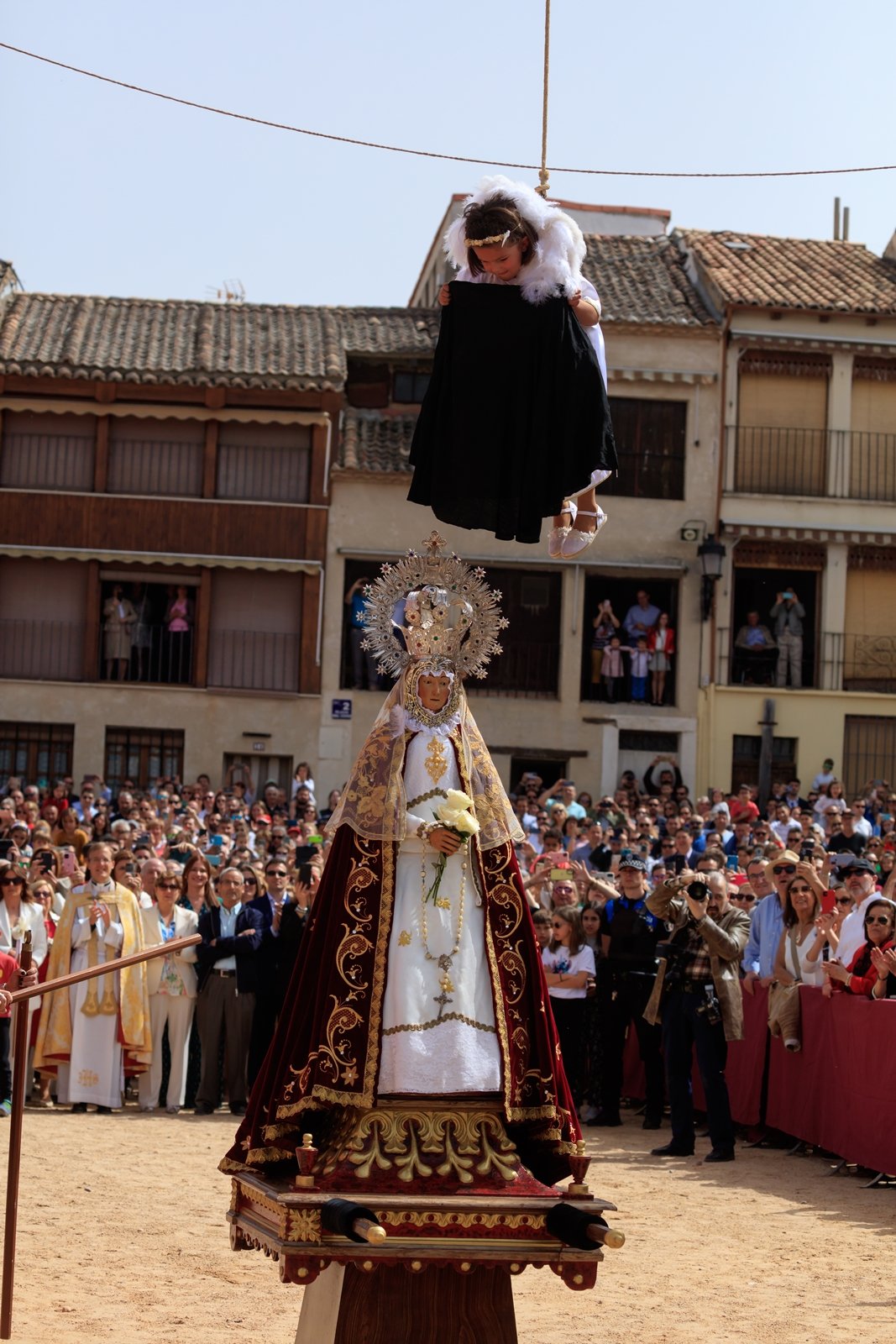 Una procesión religiosa con una imagen de la Virgen María y un niño disfrazado de ángel que vuela sobre ella. La Virgen lleva una corona y un manto decorado, y la escena es vista por una multitud en una plaza.
