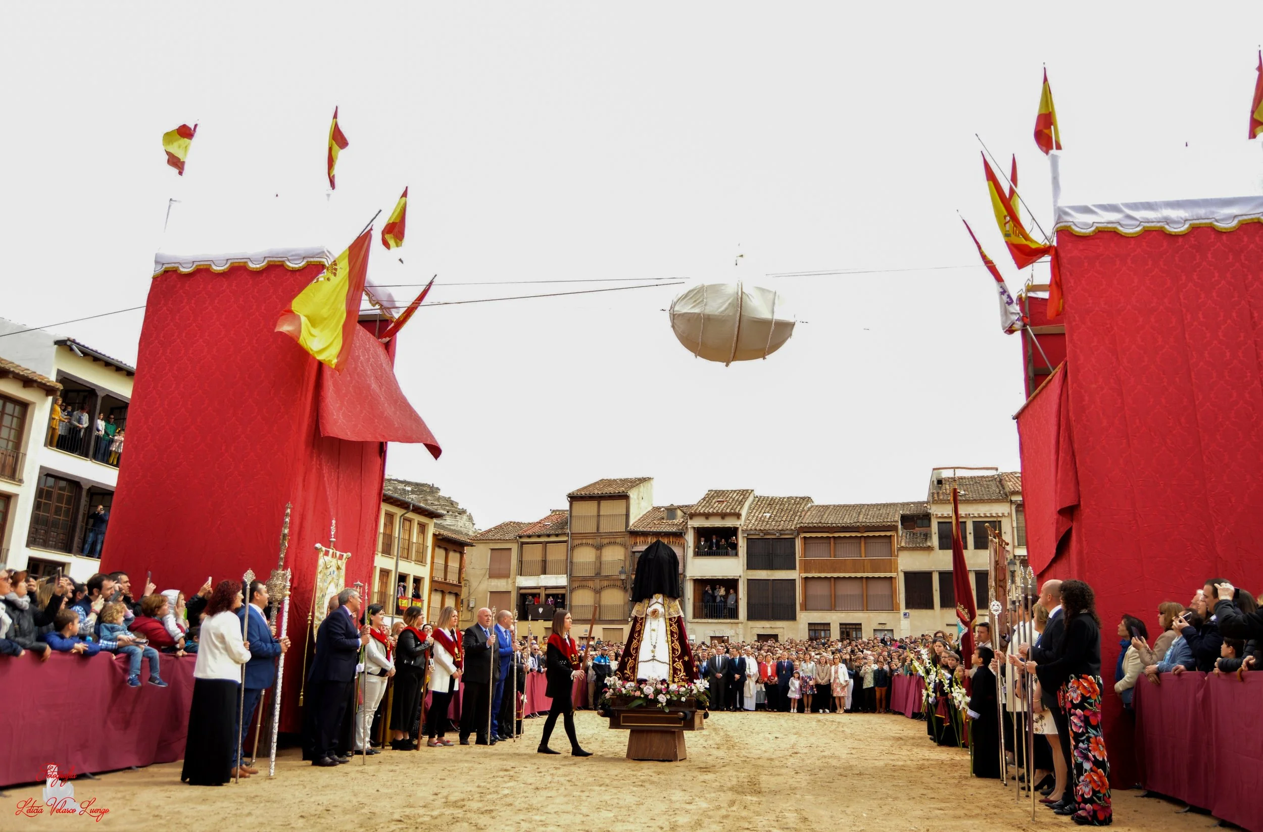 Celebración en la plaza principal con estructuras cubiertas de tela roja y banderas, frente a una multitud y una figura central decorada con flores.