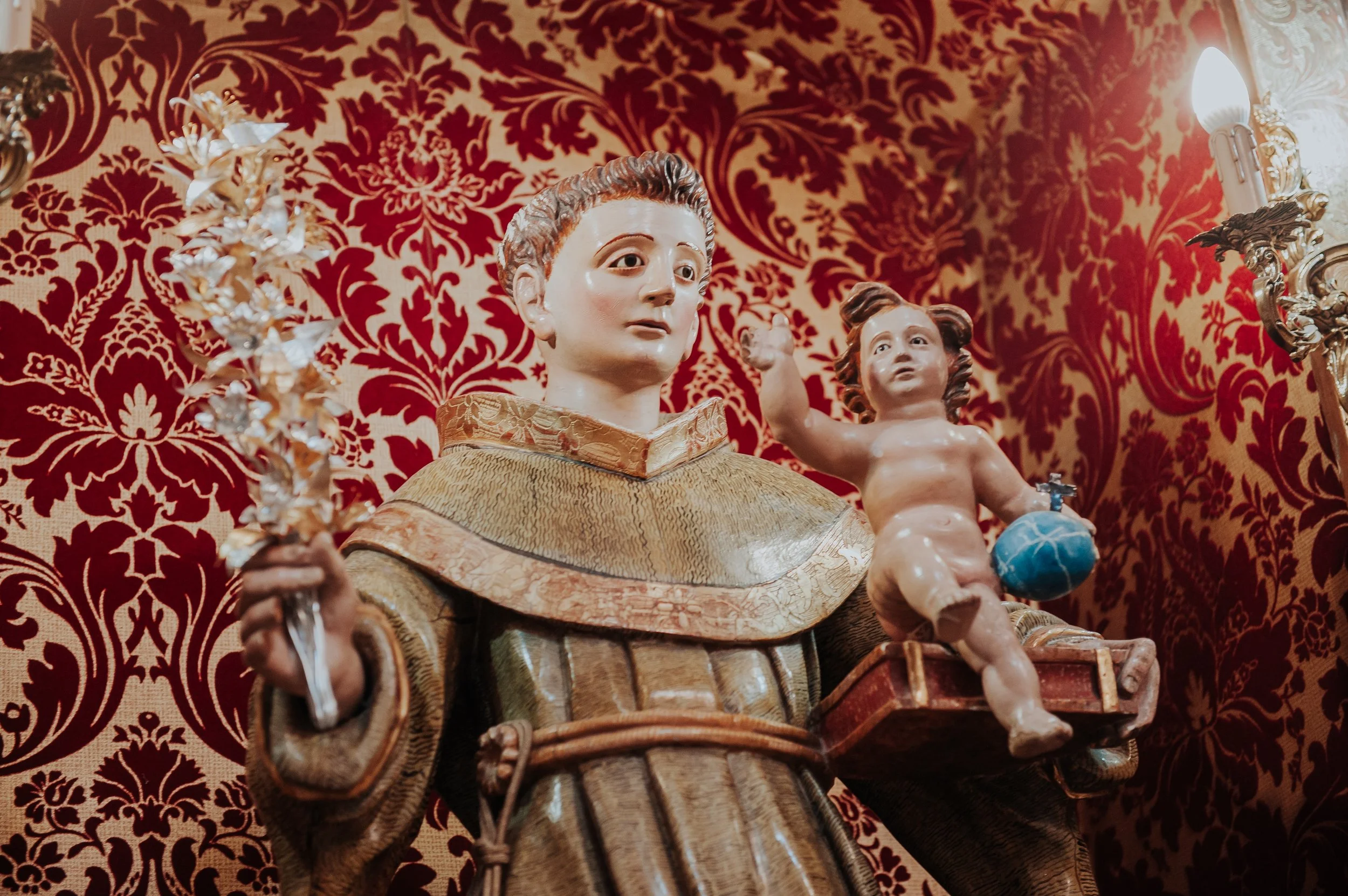 Escultura religiosa de un santo católico sosteniendo una cruz y una figura infantil con un globo azul, en un fondo de tapiz rojo con dorado.