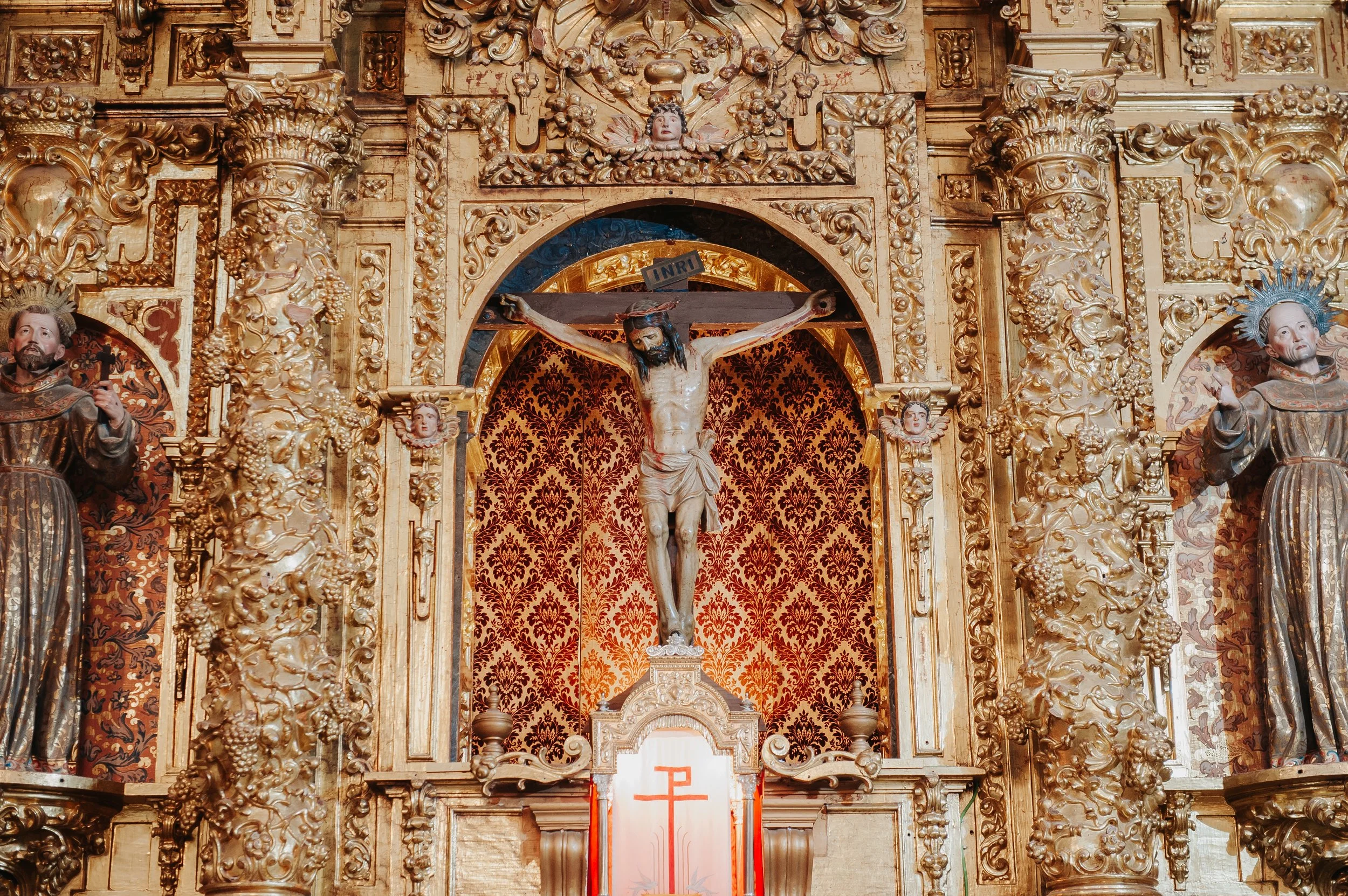 Cristo crucificado en un altar barroco dorado, con figuras de santos a los lados y fondo con tela roja y motivo dorado.