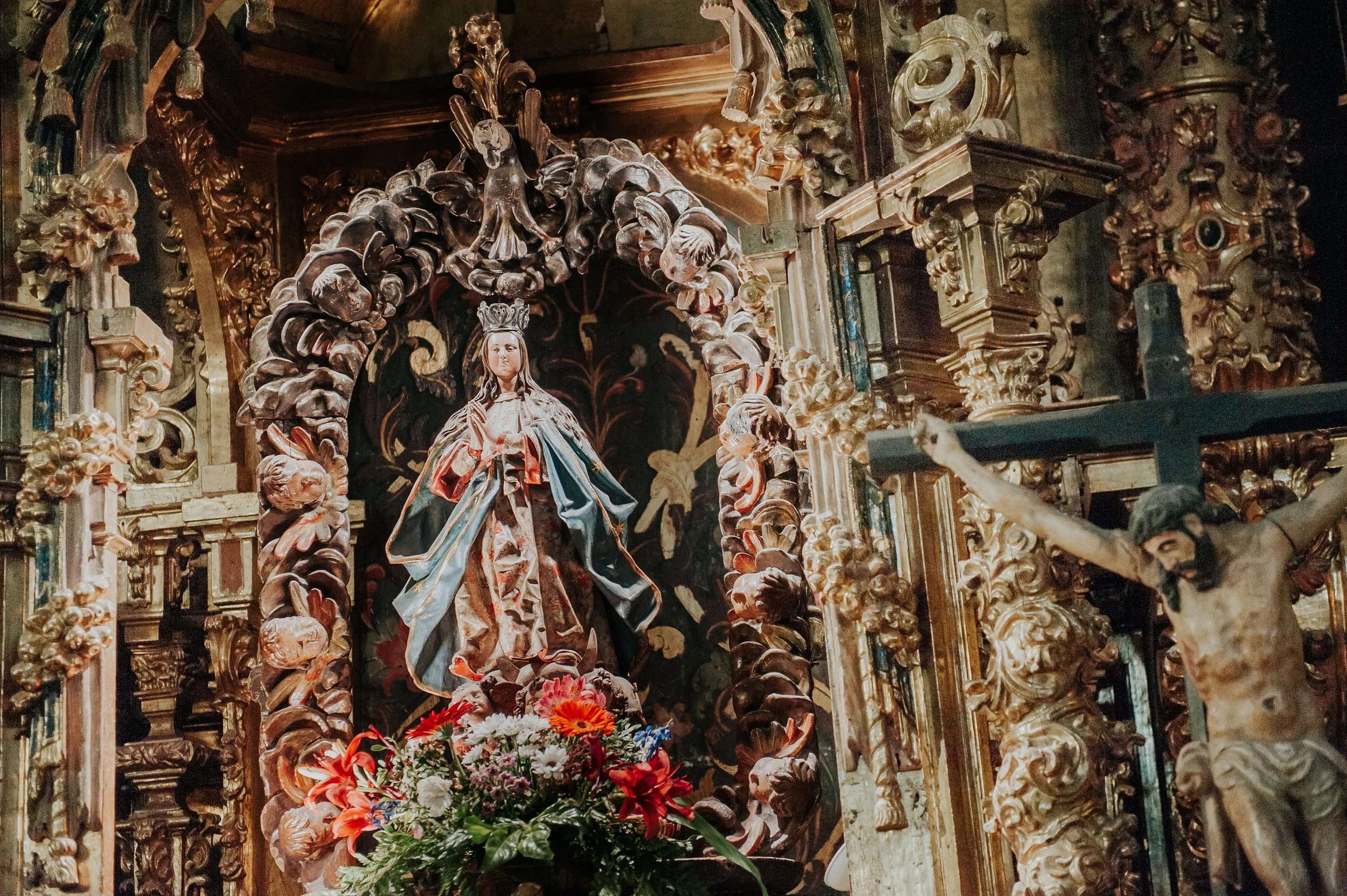 Figurillas religiosas, principalmente una Virgen con corona y un Jesús crucificado en primer plano, rodeados de detalles dorados y flores decorativas en un altar.