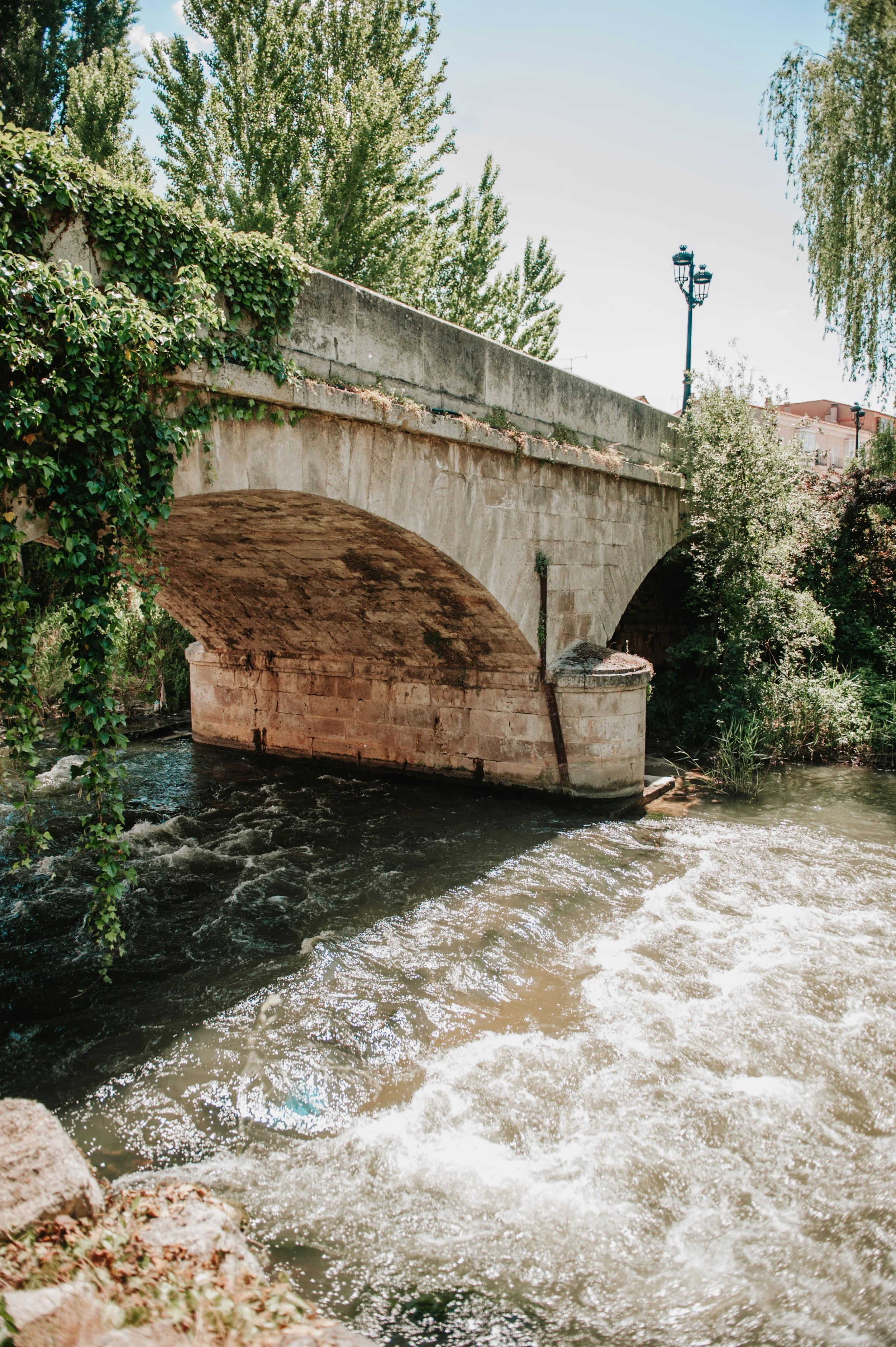 Puente de piedra sobre un río con árboles y un poste de luz en el fondo.