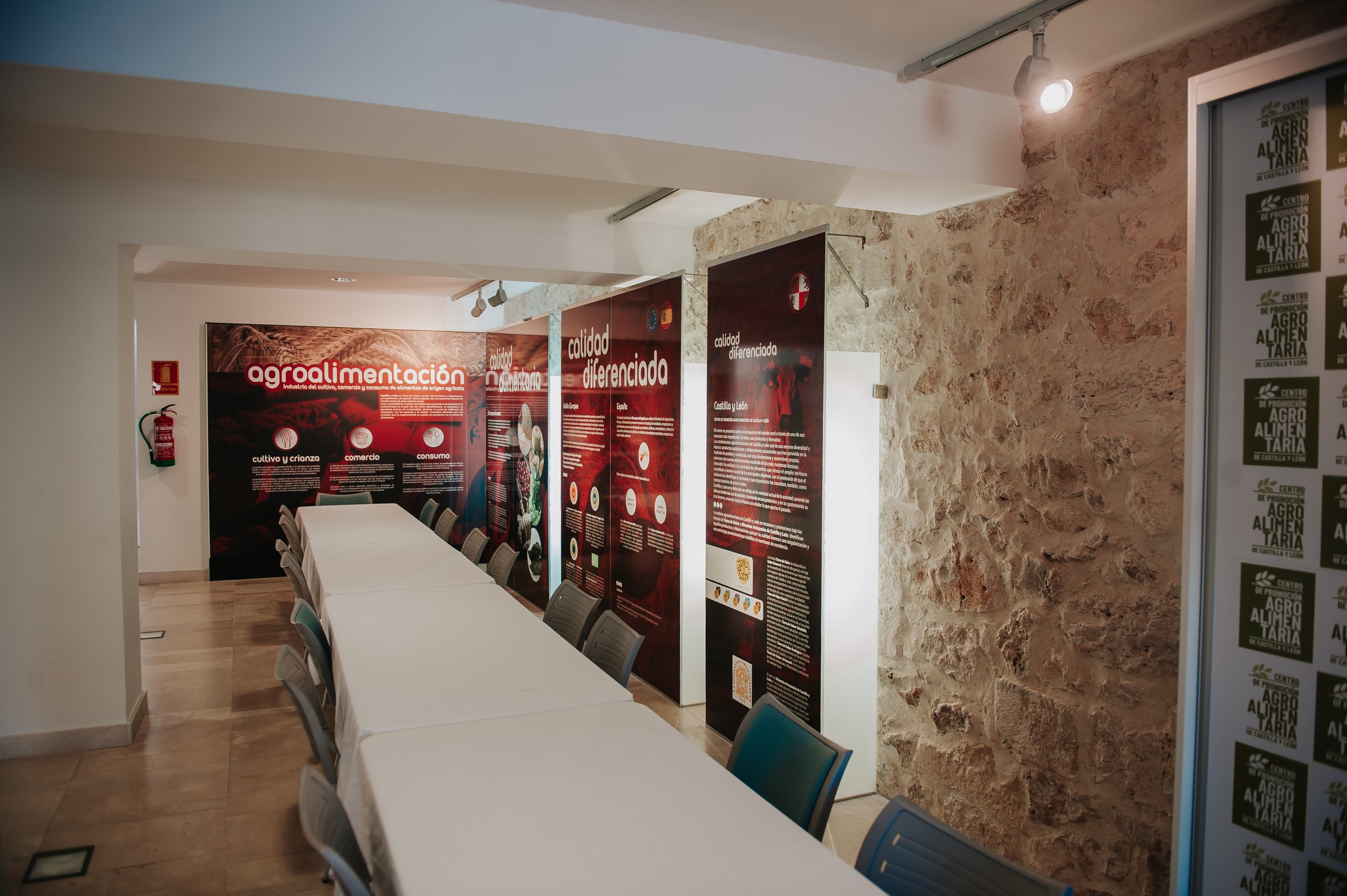 Sala de conferencia con mesas cubiertas con mantel blanco, paredes con carteles sobre agroalimentación y estructura de piedra decorativa en una de las paredes.