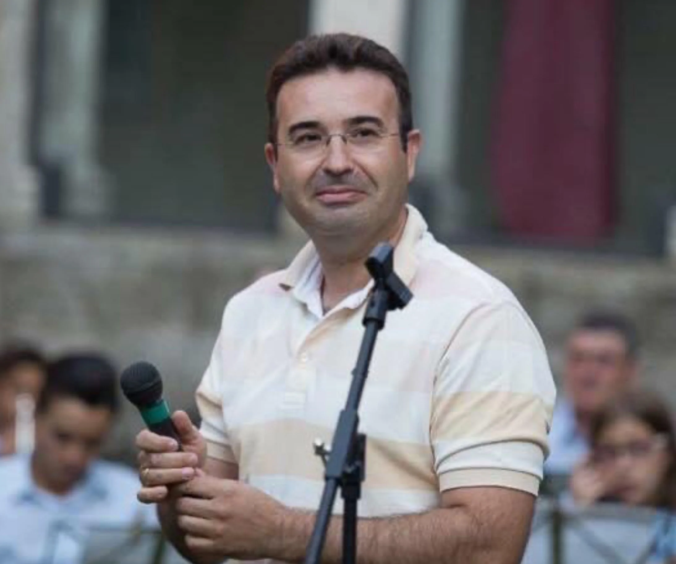 Hombre con gafas, camisa clara, sosteniendo un micrófono, en un evento público, con personas en el fondo.