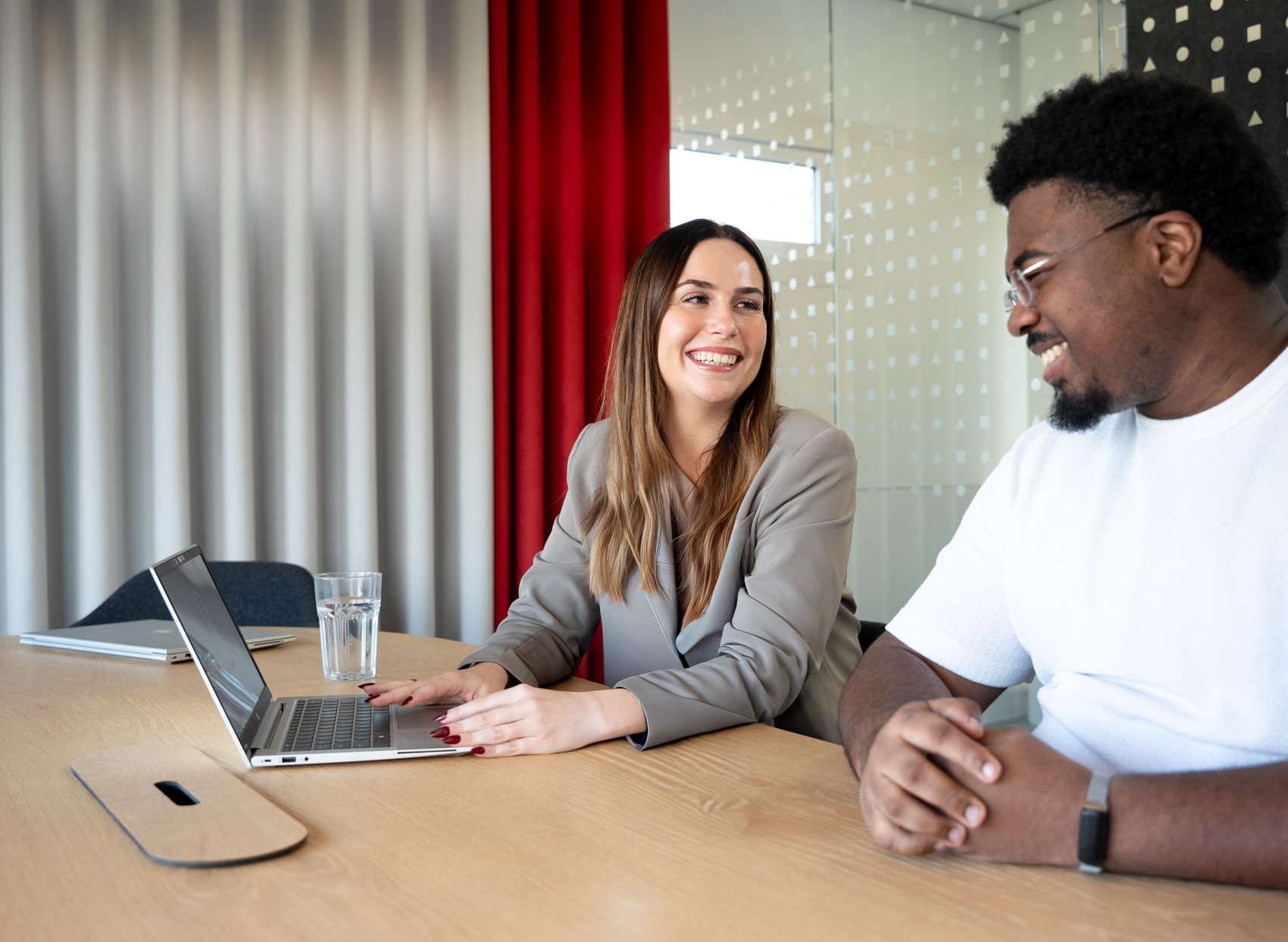 Foto van collega's lachen en praten tijdens een vergadering, met een laptop en een glas water op de tafel.