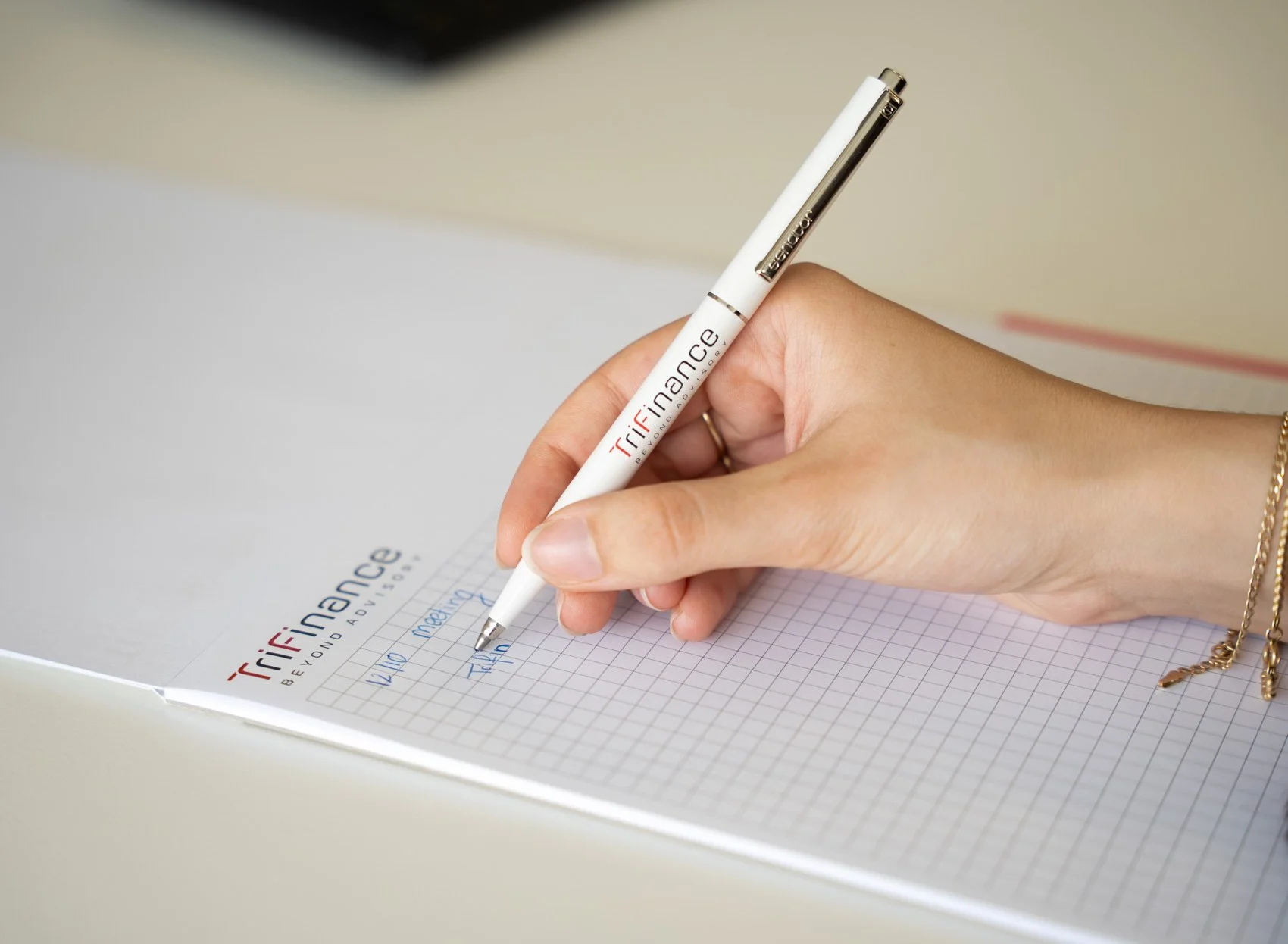 Persoon die schrijft op een geannoteerd notitieblok met een pen met het 'Trifinance Beyond Advice' logo.