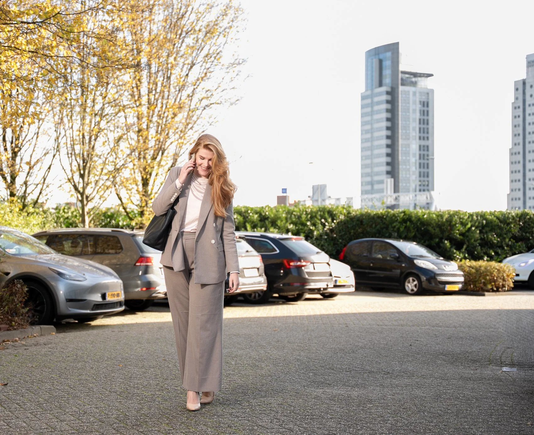 Vrouw in een grijs pak die op haar telefoon loopt in een parkeerplaats met geparkeerde auto's en hoogbouw op de achtergrond, onder een blauwe hemel.