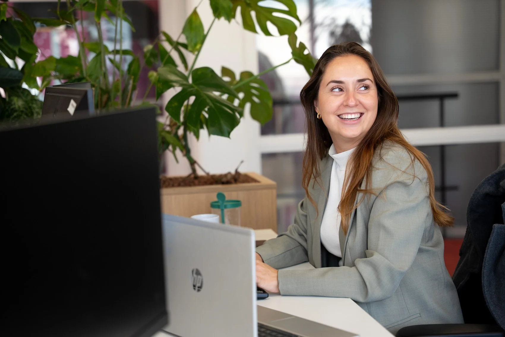 Een vrouw zit aan een kantoor bureau, lachend en kijkend naar rechts, met een computer en planten op de achtergrond.
