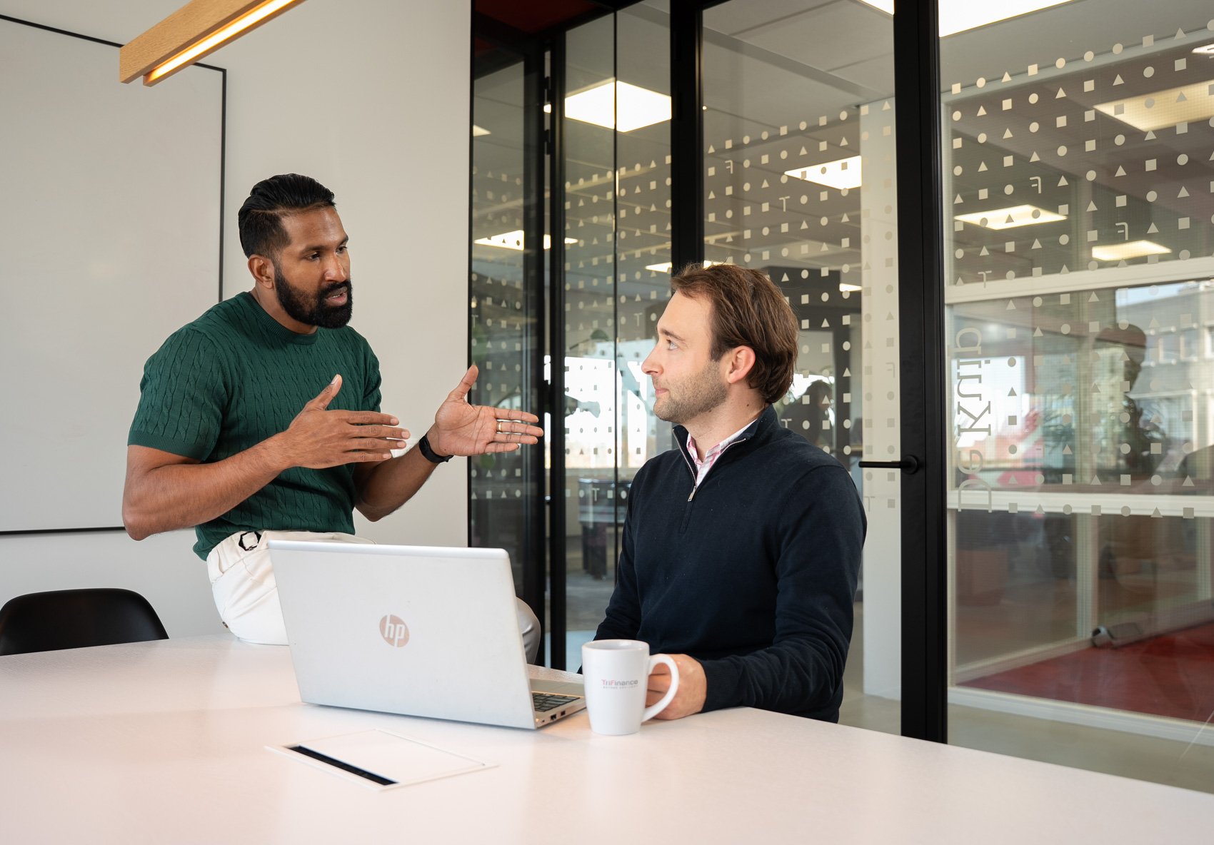 Twee mannen hebben een gesprek in een vergaderruimte met grote glazen muur en een laptop en mok op tafel.