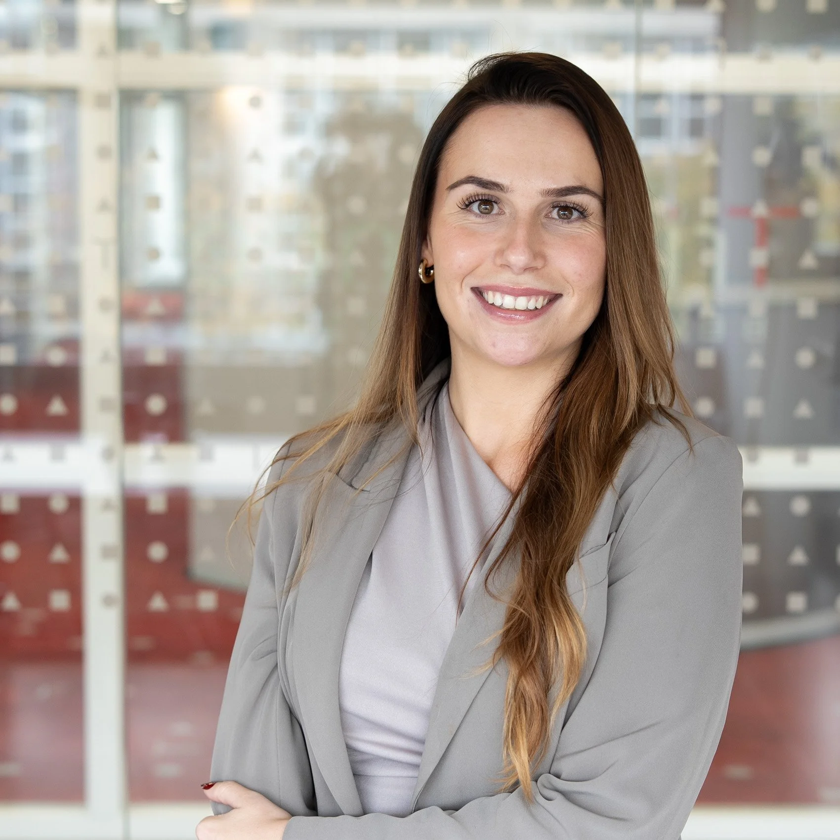 Portret van een lachende vrouw met lange, golvende, bruin haar, gekleed in een grijze jas, staand voor een glasscherm met geometrische patronen.