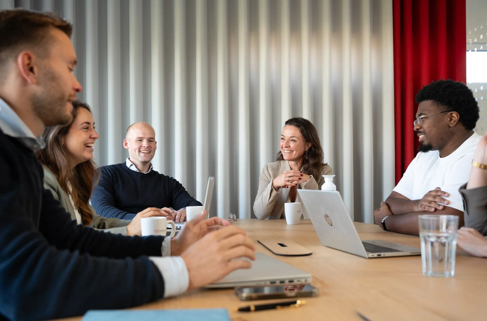 Vijf mensen zitten aan een tafel in een vergaderzaal en lachen met elkaar.