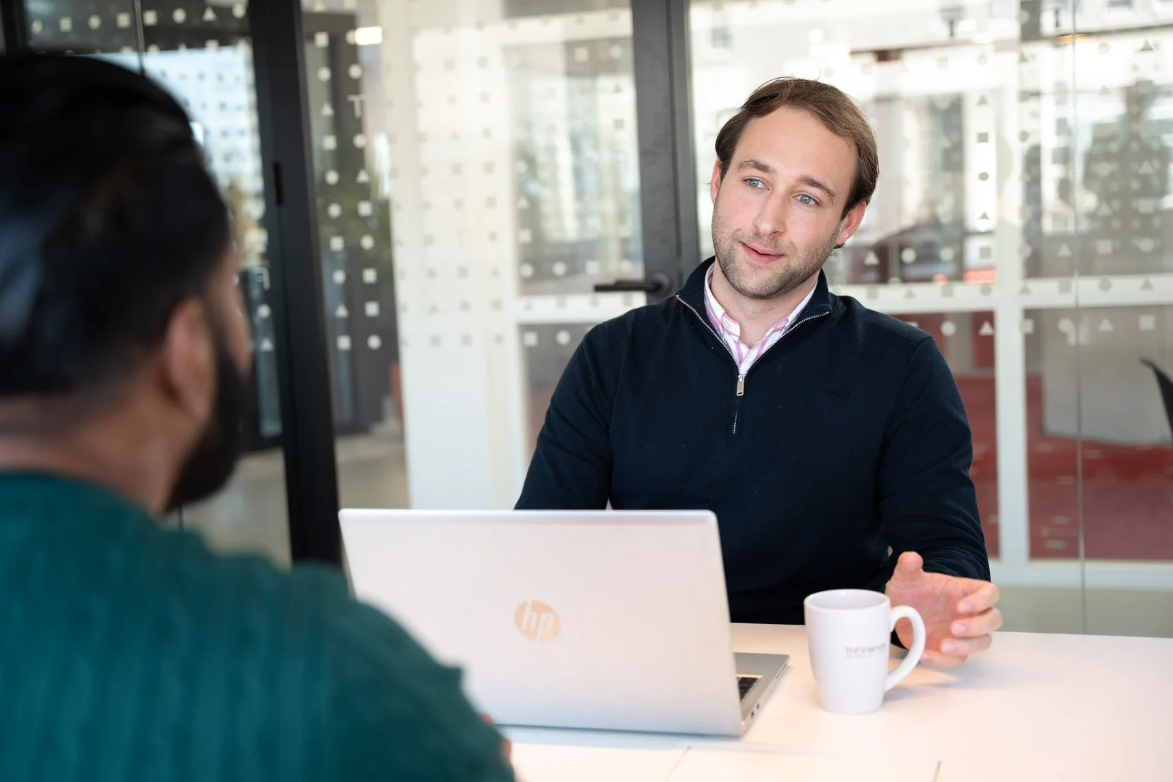 Twee mensen zitten aan een tafel, de man kijkt naar de persoon die voor hem zit en heeft een kop koffie naast zich, er is een laptop op de tafel.