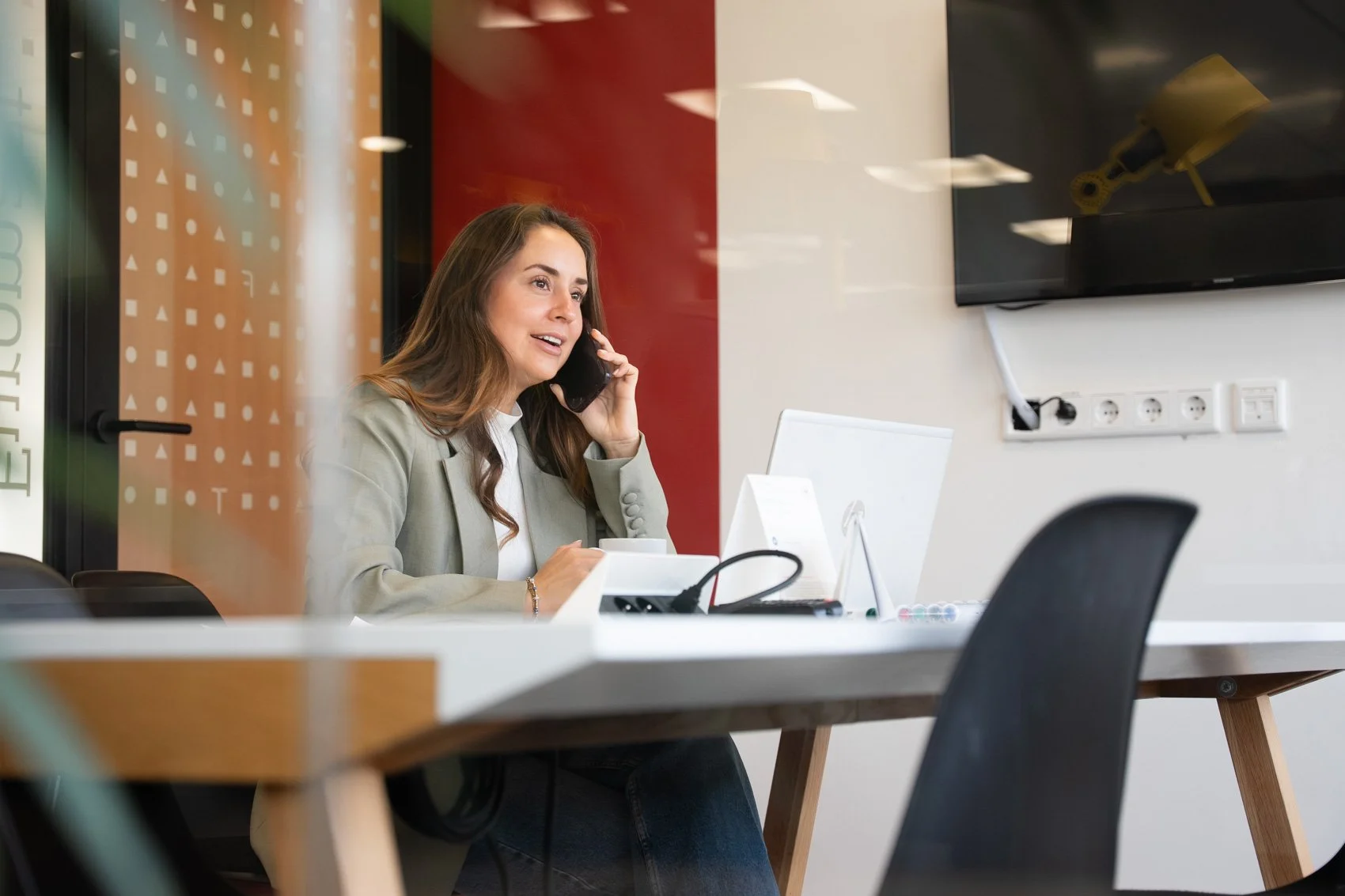 Vrouw zit aan een tafel in een kantoorruimte, belt met een mobieltje, naast een open laptop.