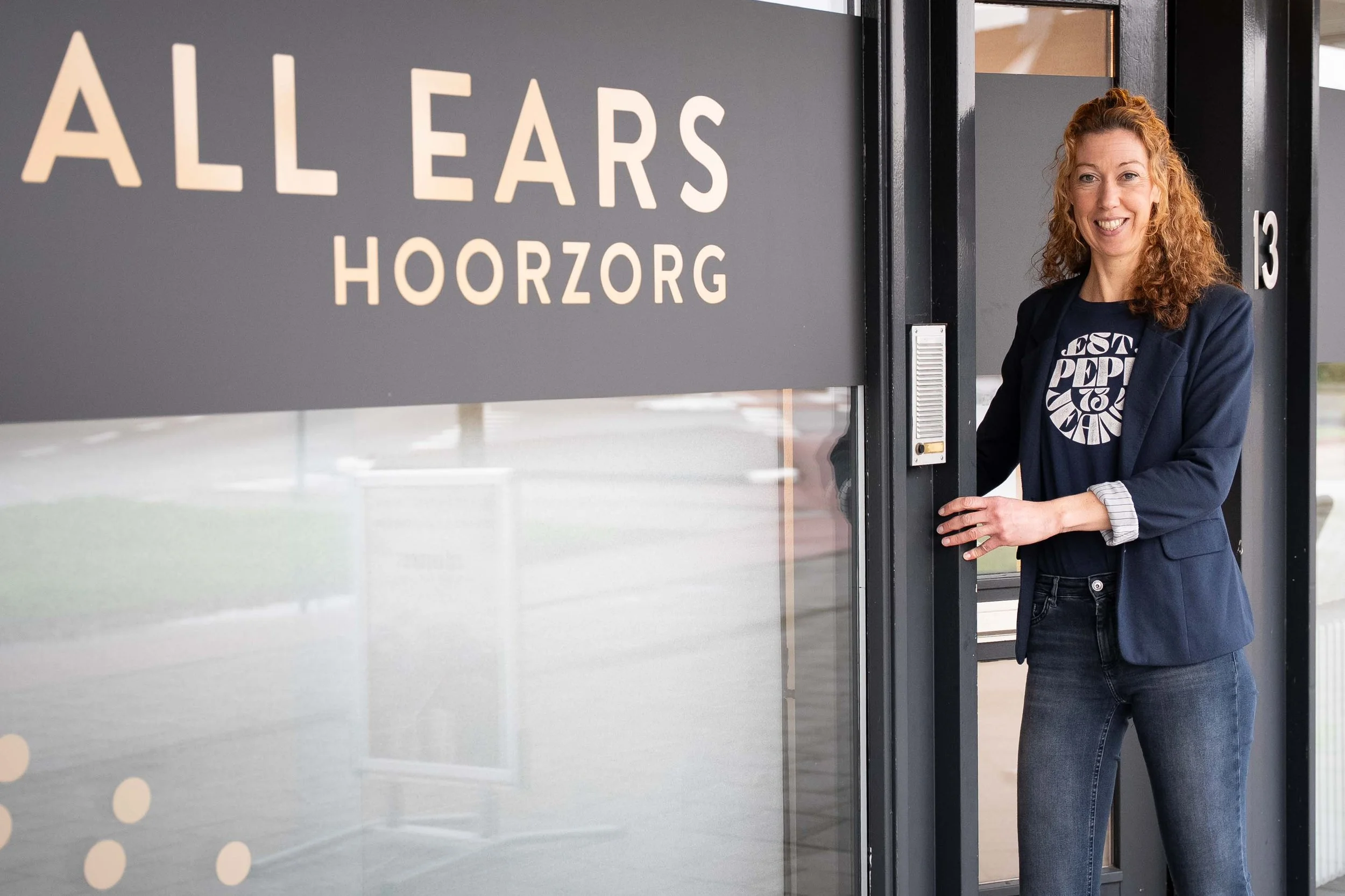 Vrouw met rood haar die de deur opent van een gebouw met een bord waarop staat 'ALL EARS HOORZORG' tijdens zakelijke fotoshoot door Studio Brenda bedrijfsfotografie