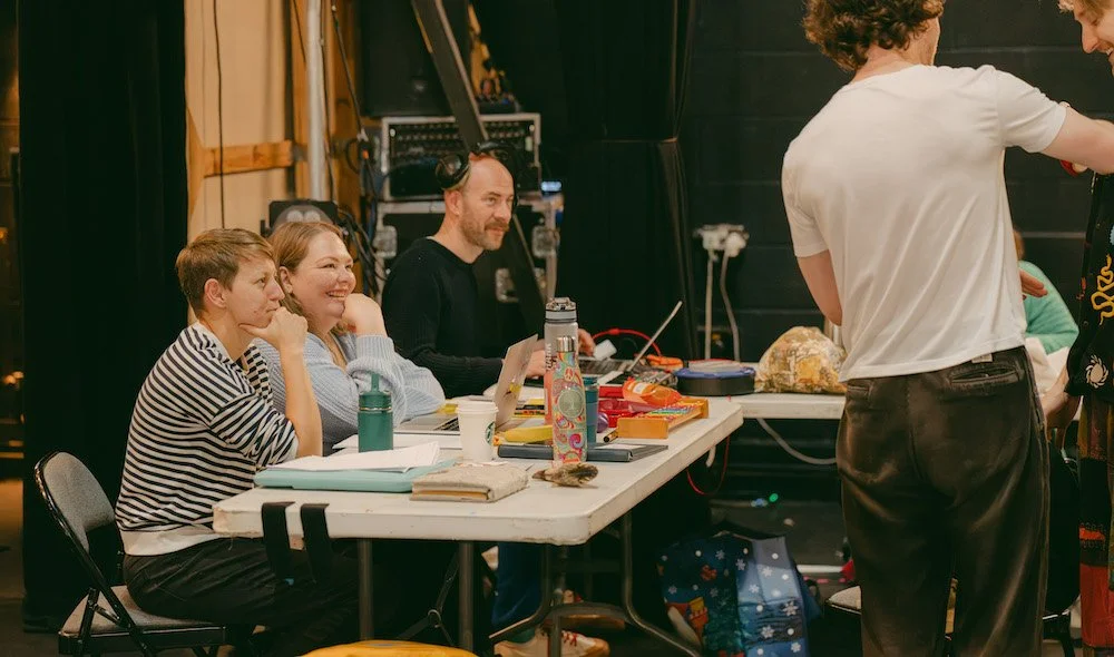 Three people sit at a pale grey table in a theatre space, smiling as they watch something off-camera, with sets and equipment behind them.