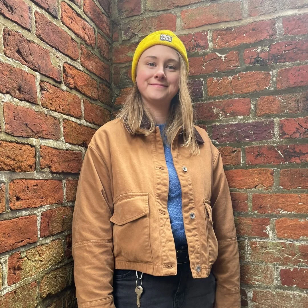 A person with shoulder length blonde hair stands in front of a brick wall, smiles at the camera and wears a yellow beanie, a tan jacket, and a blue sweater.