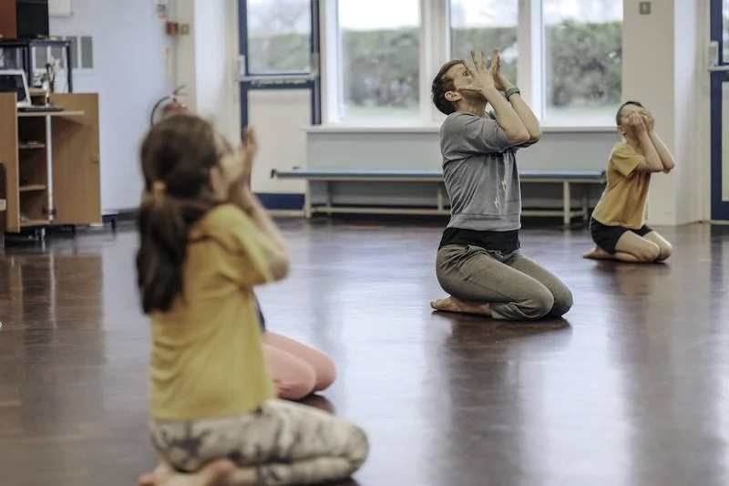 A child in yellow and an adult in grey kneel in a school hall with hands raised to their faces, fingers outstretched, looking upward.